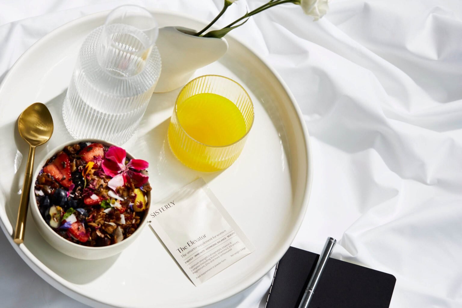 All-in-one supplement blend for women by Sisterly supplement brand for women, shown with a glass of juice, granola bowl with fruit, and a supplement sachet on a tray