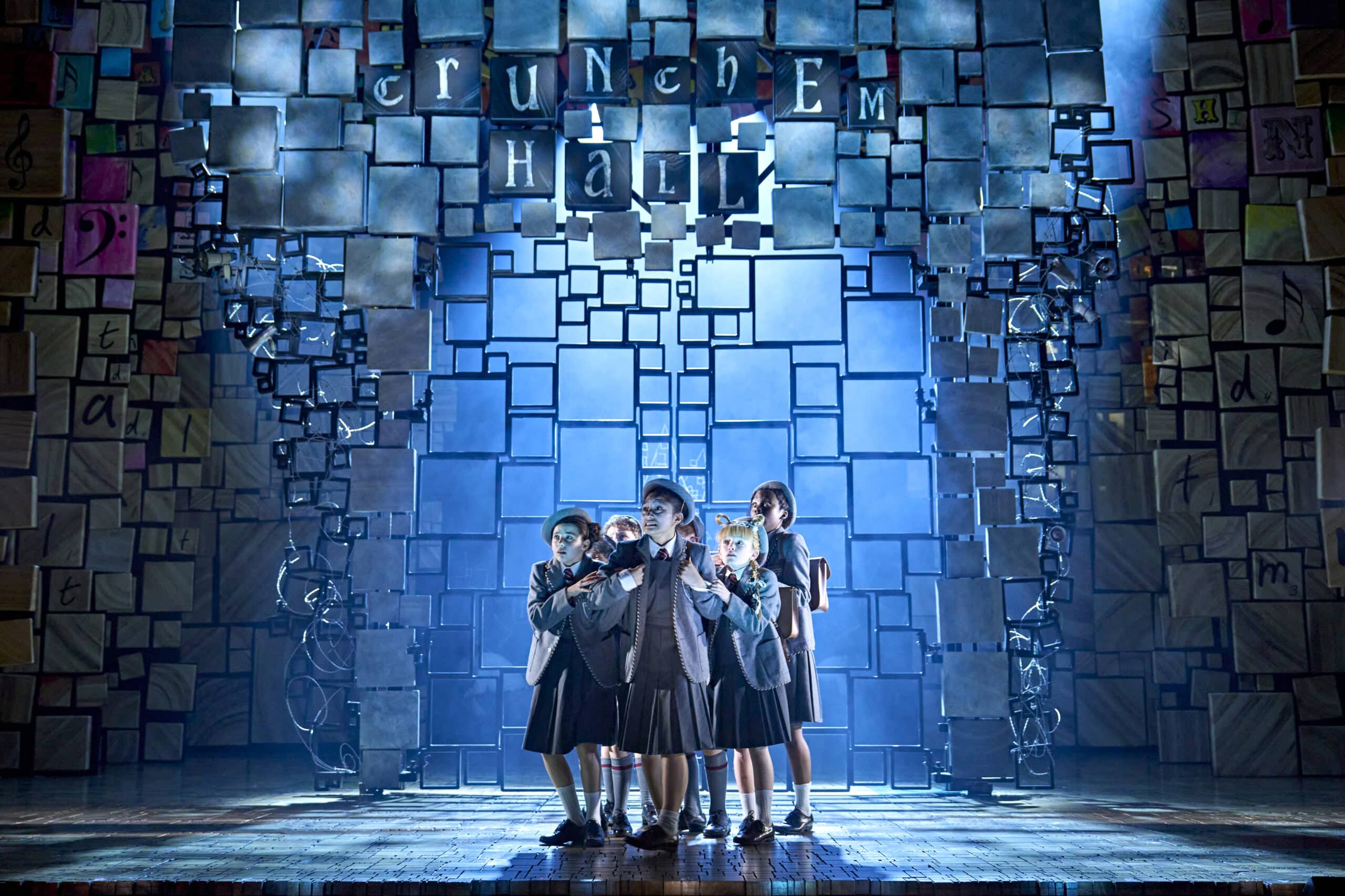 children on stage for matilda the musical. dressed in grey school uniforms and huddled together under blue light