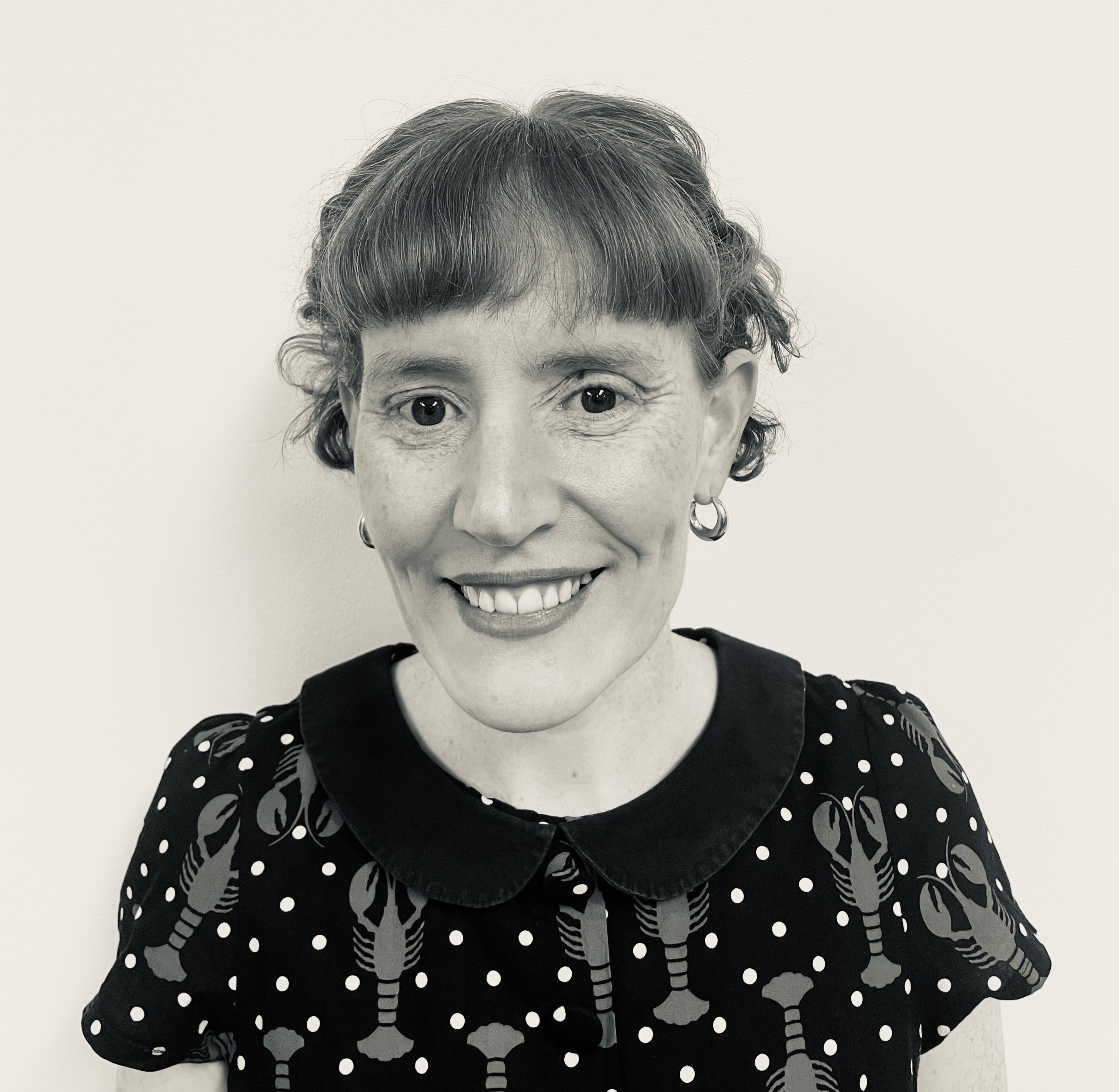 Black-and-white portrait of author Rosa Campbell smiling at the camera while wearing a patterned dress with a dark collar, photographed for The Women's Journal interview for The Book that Taught the World to Orgasm and then Disappeared review