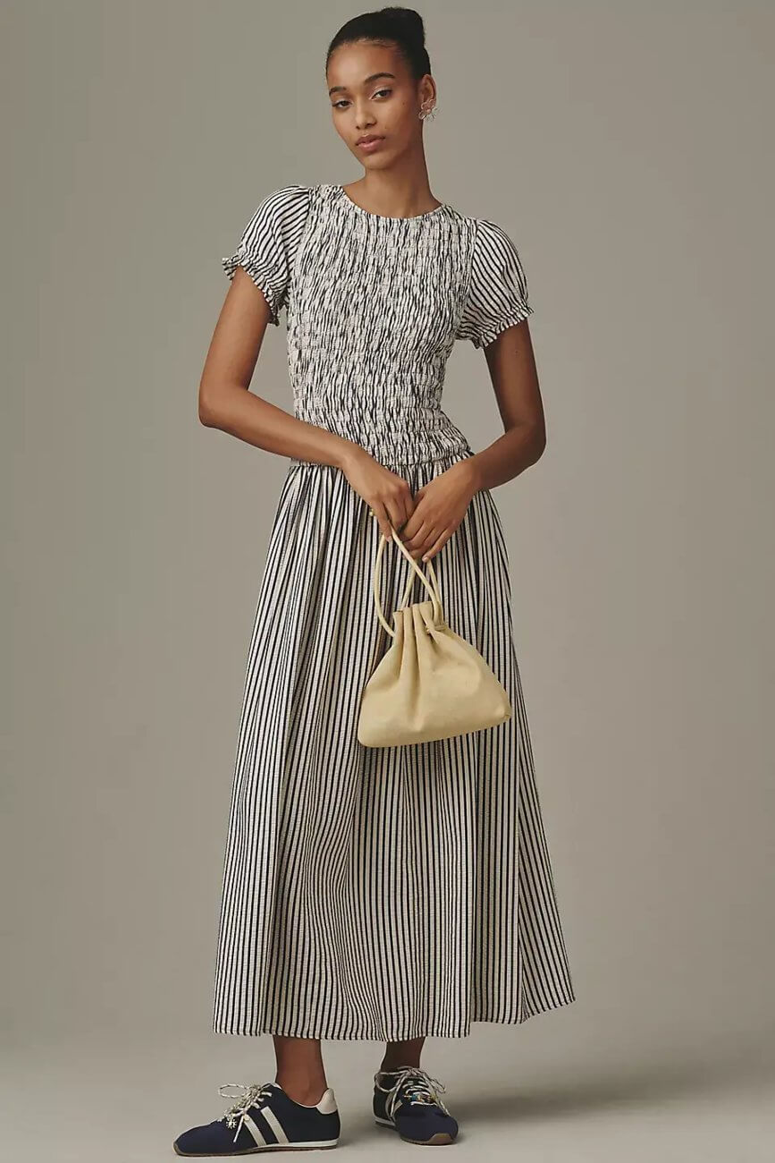 Model wearing Anthropologie Maeve Smoked Short Sleeve Maxi Dress, a striped short sleeve maxi dress with a gathered bodice, holding a small bag