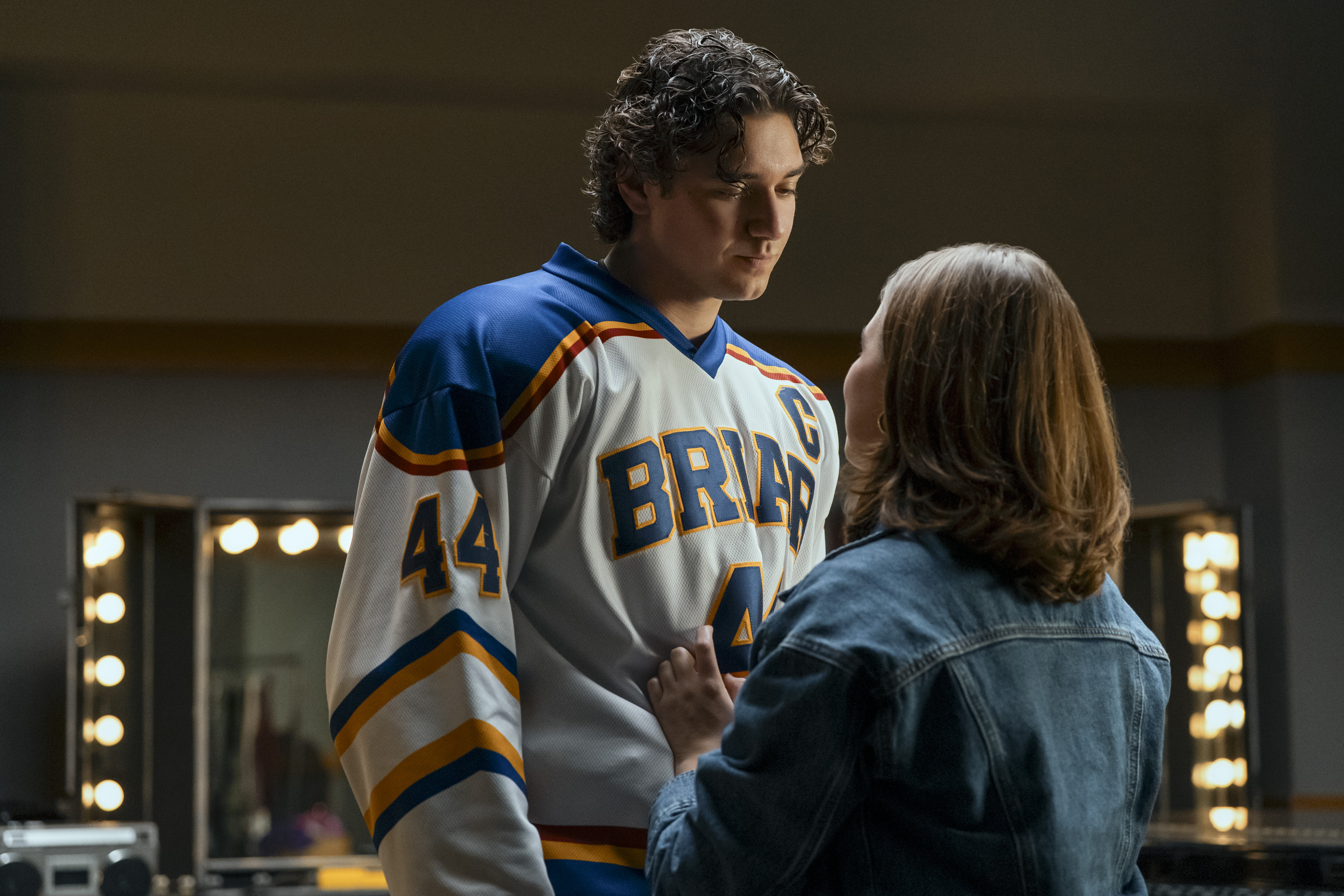 Belmont Cameli as Garrett, in a white 'Briar' hockey jersey, looks down thoughtfully at Ella Bright as Hannah, who wears a denim jacket and has her hand on his chest in a scene from 'Off Campus'.