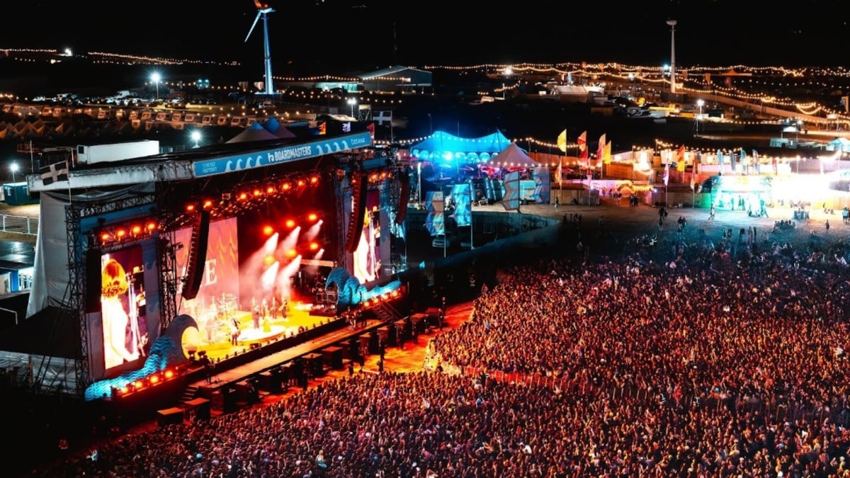 A high-angle nighttime view of a massive crowd packed in front of the brightly lit Main Stage at Boardmasters Festival, featuring wave-shaped stage decorations, large video screens, and a wind turbine visible in the illuminated background.