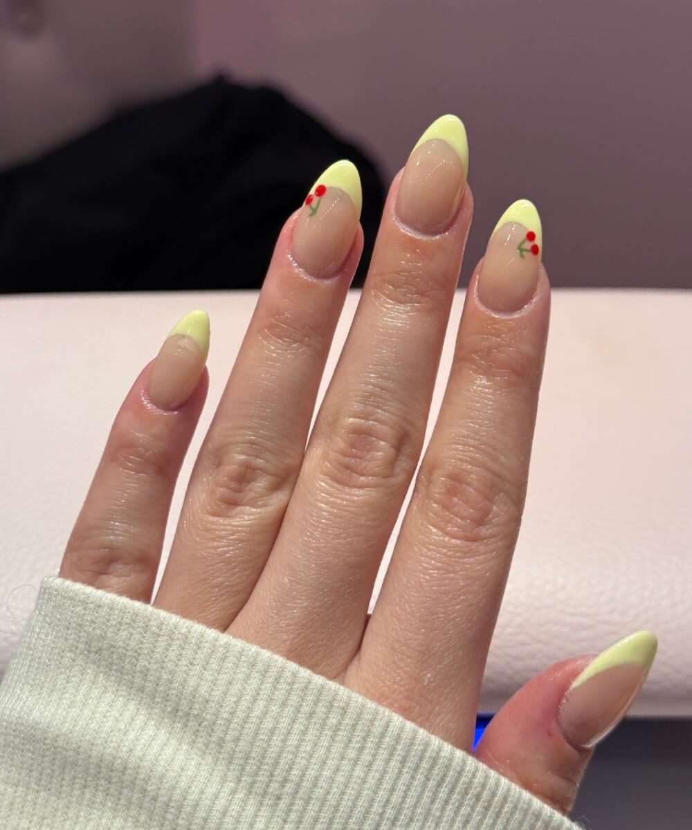 Butter yellow French tip nails with a glossy nude base and small cherry nail art on almond-shaped nails