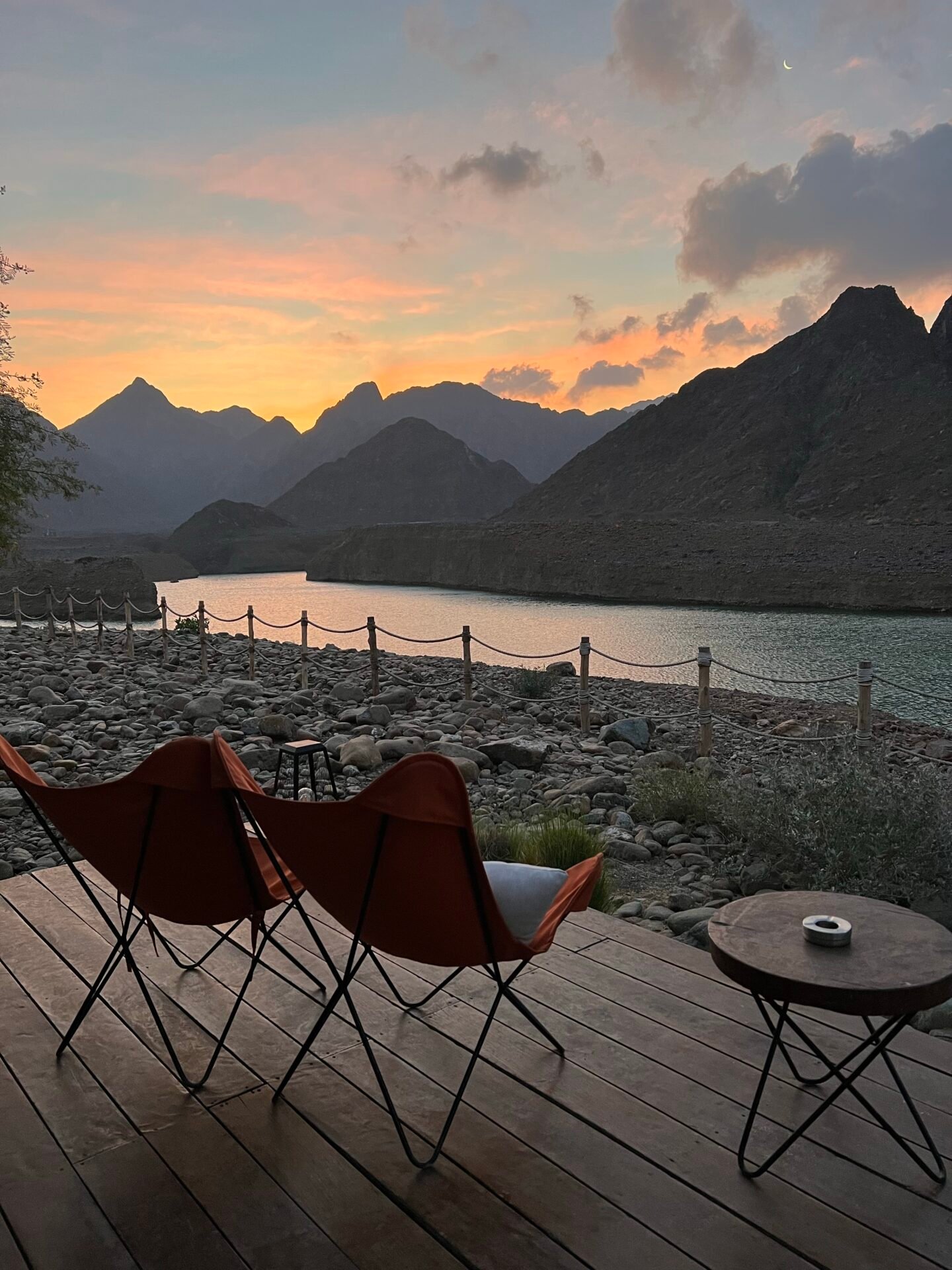 Two orange folding chairs and a small round table on a wooden deck overlooking a rocky shoreline, a calm lake, and jagged mountains at sunset representing the idea of a wellness retreat