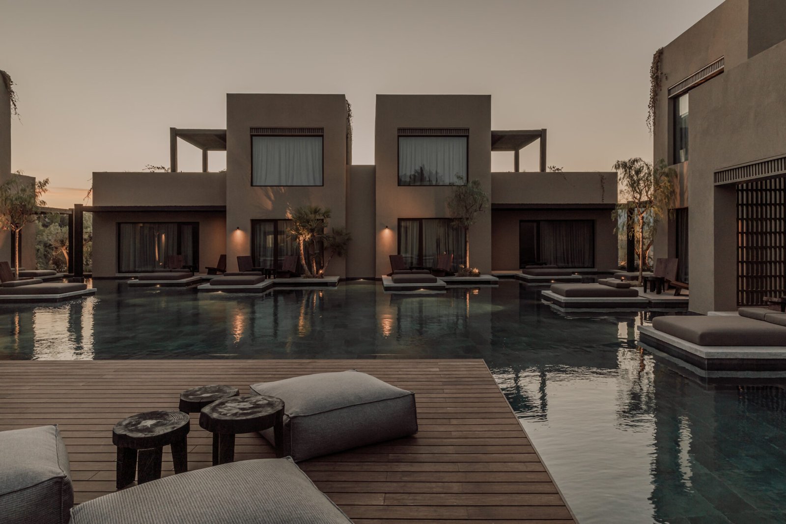 Casa Cook Samos, a modern luxury resort with minimalist beige buildings, a dark swimming pool, wooden deck, grey floor cushions, and small wooden side tables at dusk
