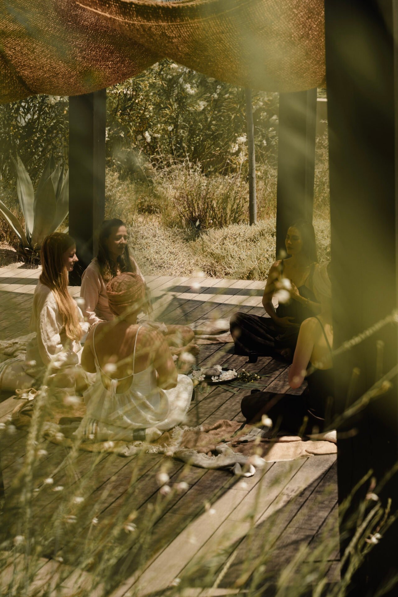 Four women from Shakti Alchemy sit in a circle on a wooden deck under a woven shade canopy, surrounded by natural greenery, engaged in a quiet outdoor gathering - best women-only retreats