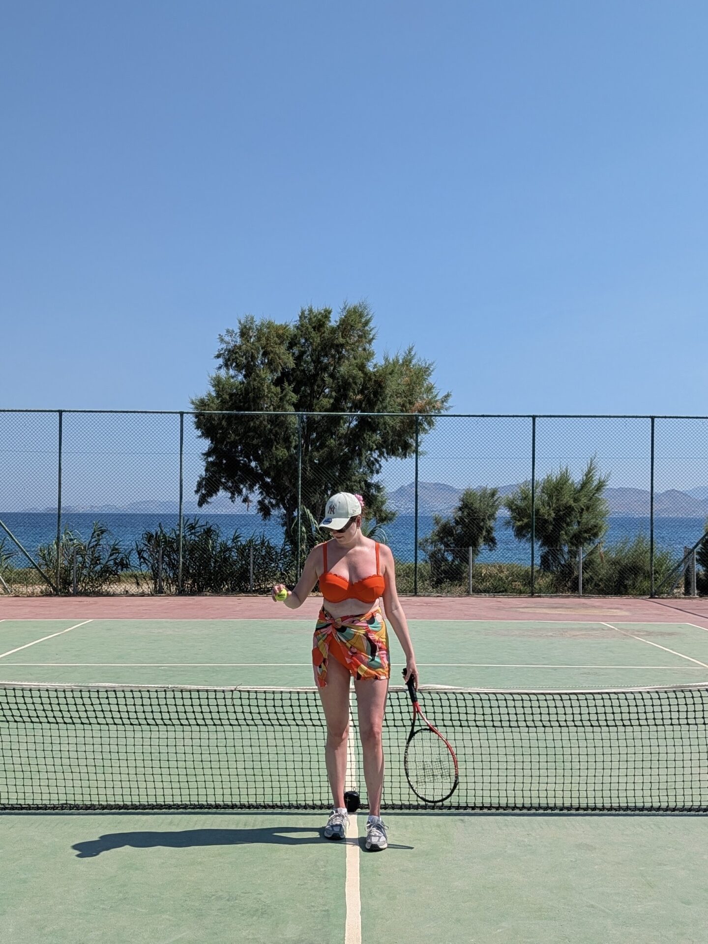 A woman stands at the net on an outdoor tennis court, wearing a bright orange JD Williams bikini top for DD+ boobs, a colourful patterned sarong, a baseball cap, and trainers. She is holding a tennis racket and a tennis ball, with a view of green trees and the blue sea behind the court's chain-link fence.