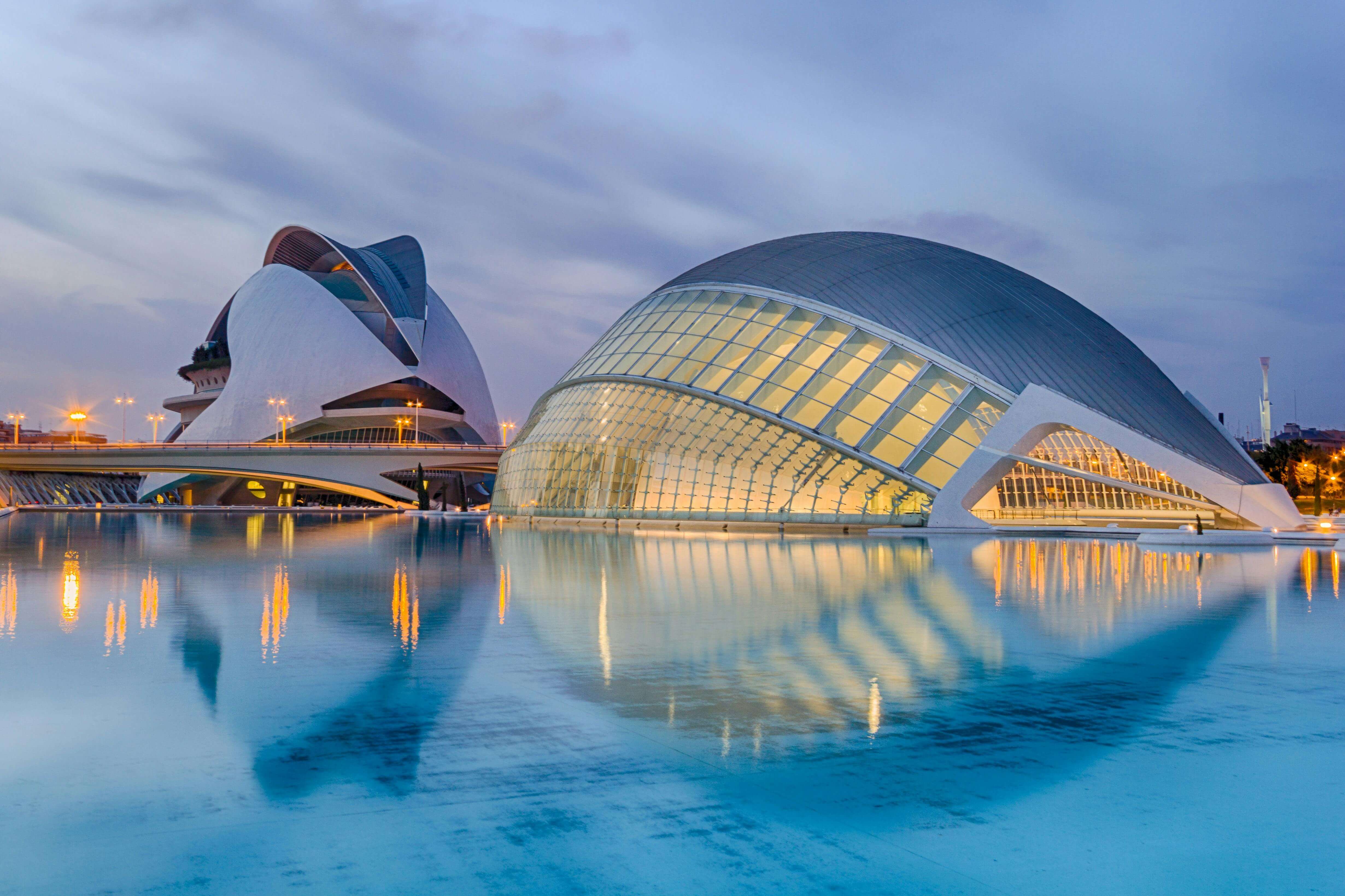 Futuristic buildings of the City of Arts and Sciences in Valencia, Spain, illuminated at dusk and reflected in a calm blue pool, featuring curved glass structures and modern architectural design.