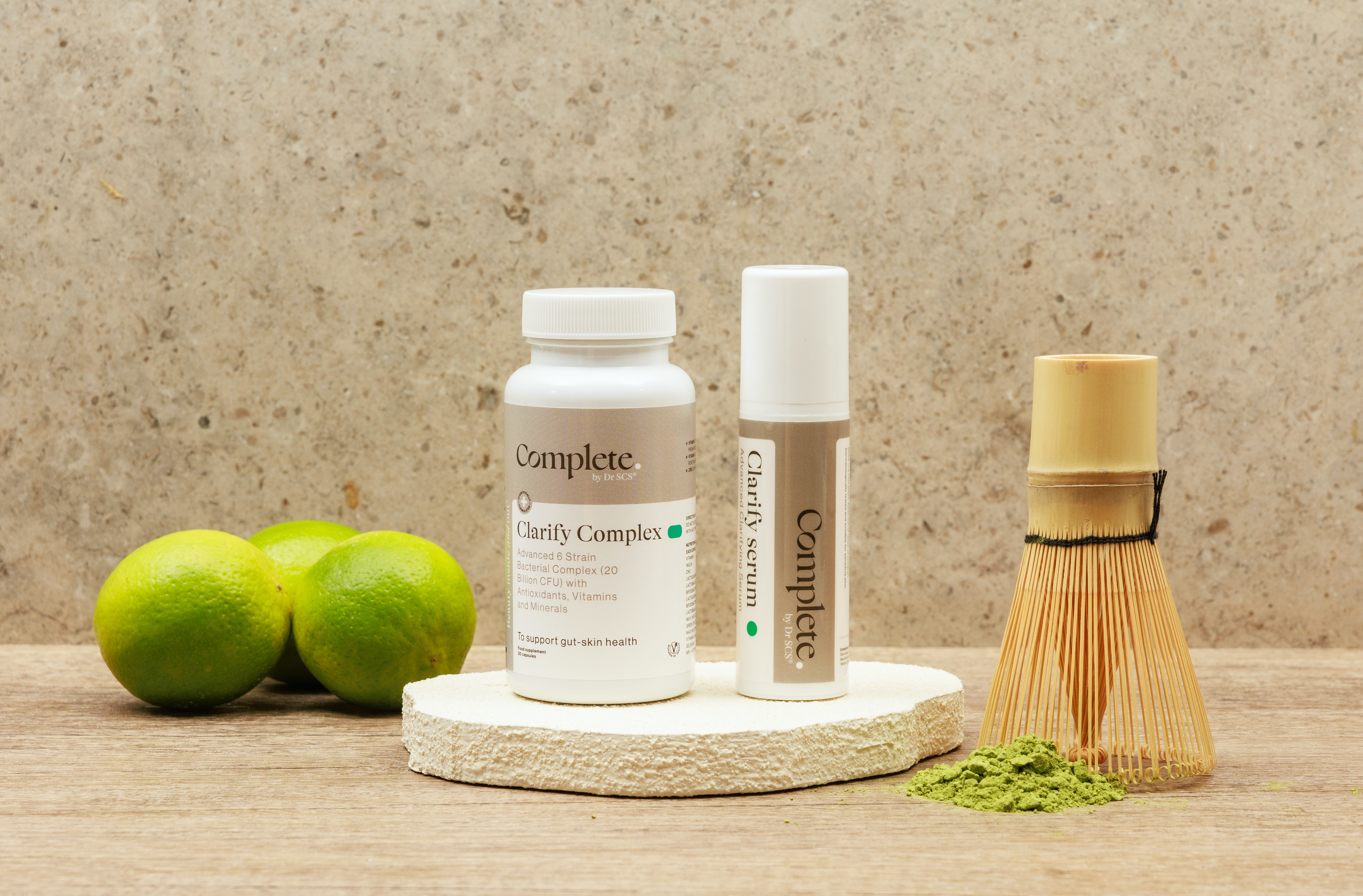 a pot of supplements and a serum in grey and white packaging positioned next to some limes and a green tea matcha whisk