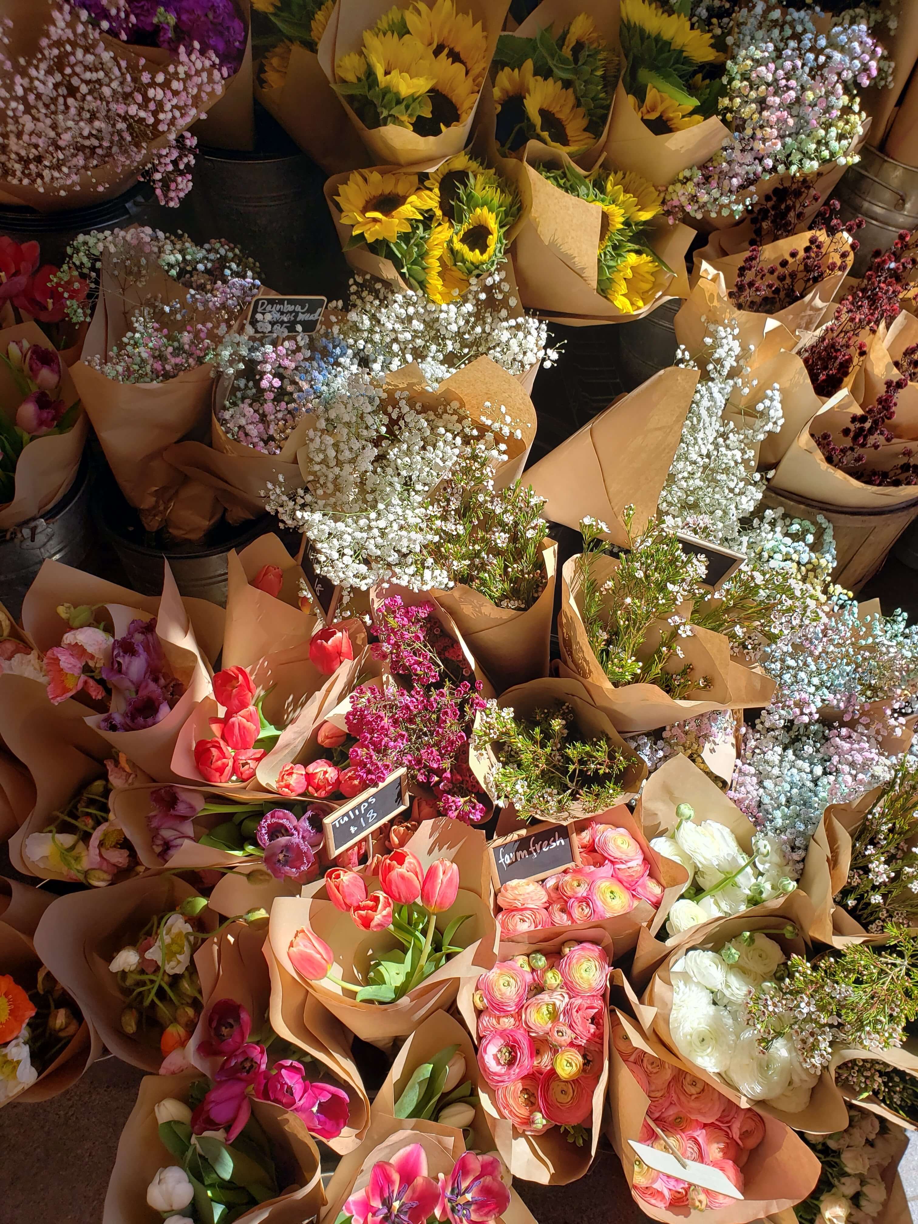 Colourful spring flower bouquets including tulips, ranunculus, baby’s breath and sunflowers wrapped in brown paper and displayed in buckets at a flower market for the edit of the best flower delivery services this Mother's Day