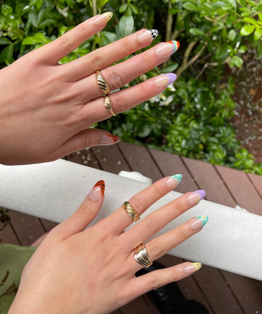Almond-shaped spring nails with colourful French tips in pastel shades, featuring mix-and-match designs including lilac, green, orange and animal print details on a glossy nude base