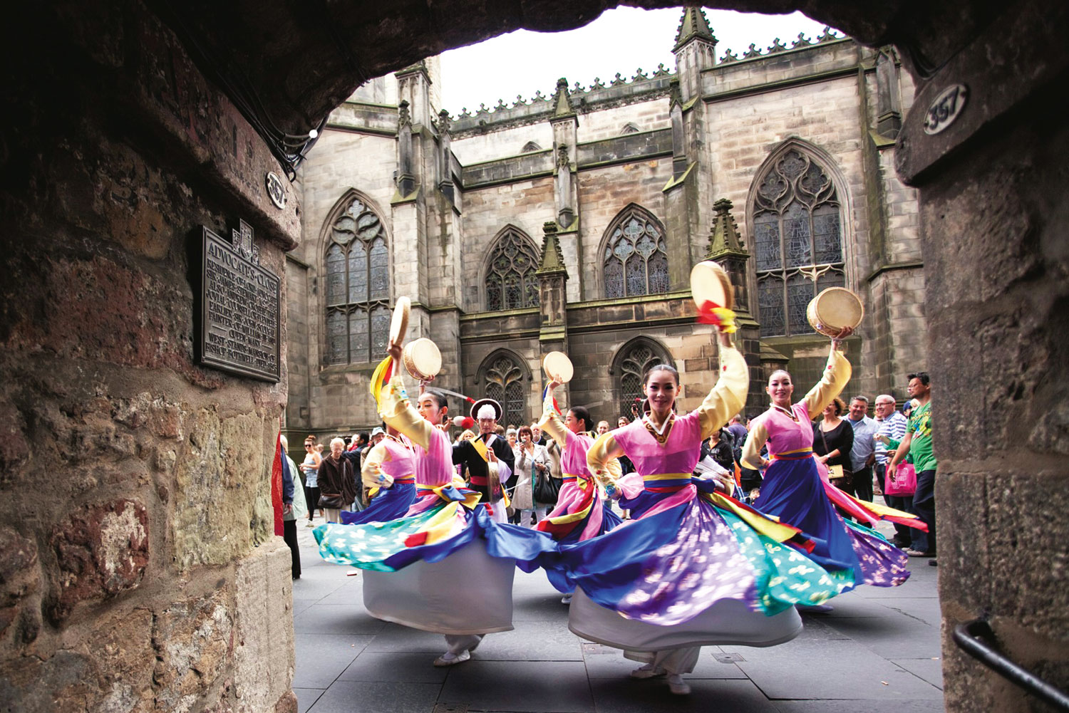 Traditional Korean dancers spin in vibrant dresses and hold drums while performing outdoors for a crowd in front of a historic stone building at the Edinburgh Festival Fringe.