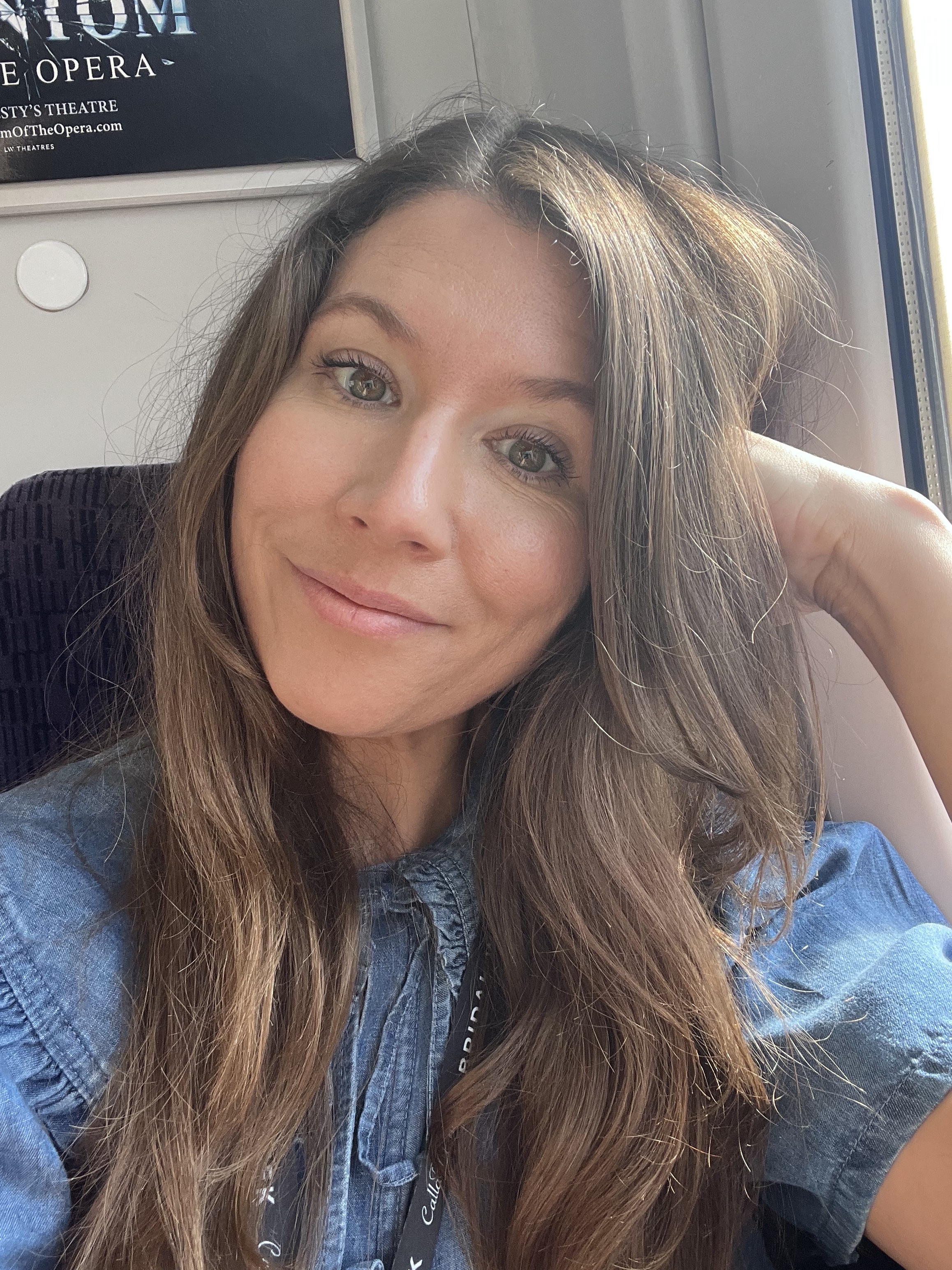 The Women's Journal journalist Danielle Wilkins with long brown hair smiles softly while resting her head on her hand, sitting by a sunlit window on the train. She’s wearing a blue top and glowing after reviewing the best adult acne treatments and products that actually work