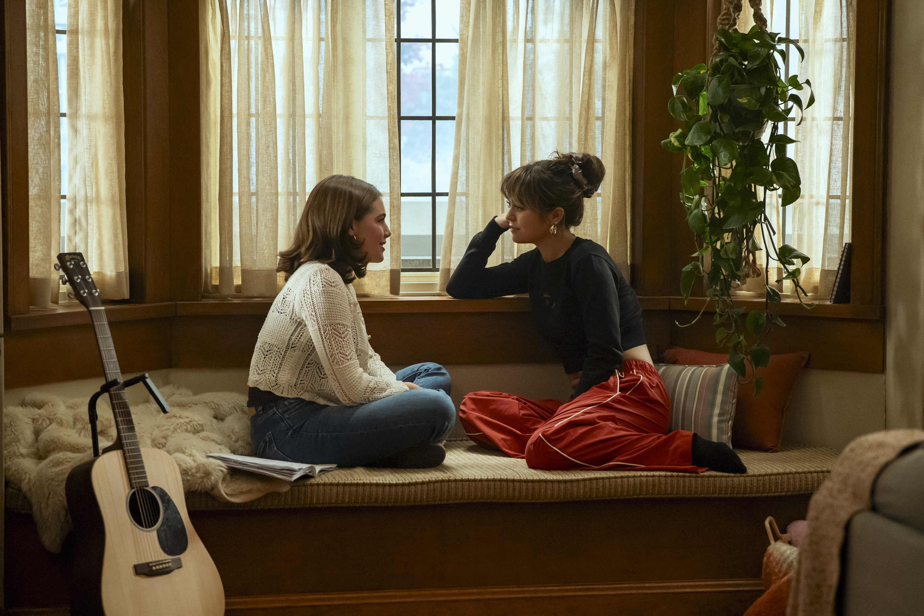 Ella Bright as Hannah and Mika Abdalla as Allie are sitting cross-legged on a cushioned window seat, facing each other in conversation in a scene from 'Off Campus'.