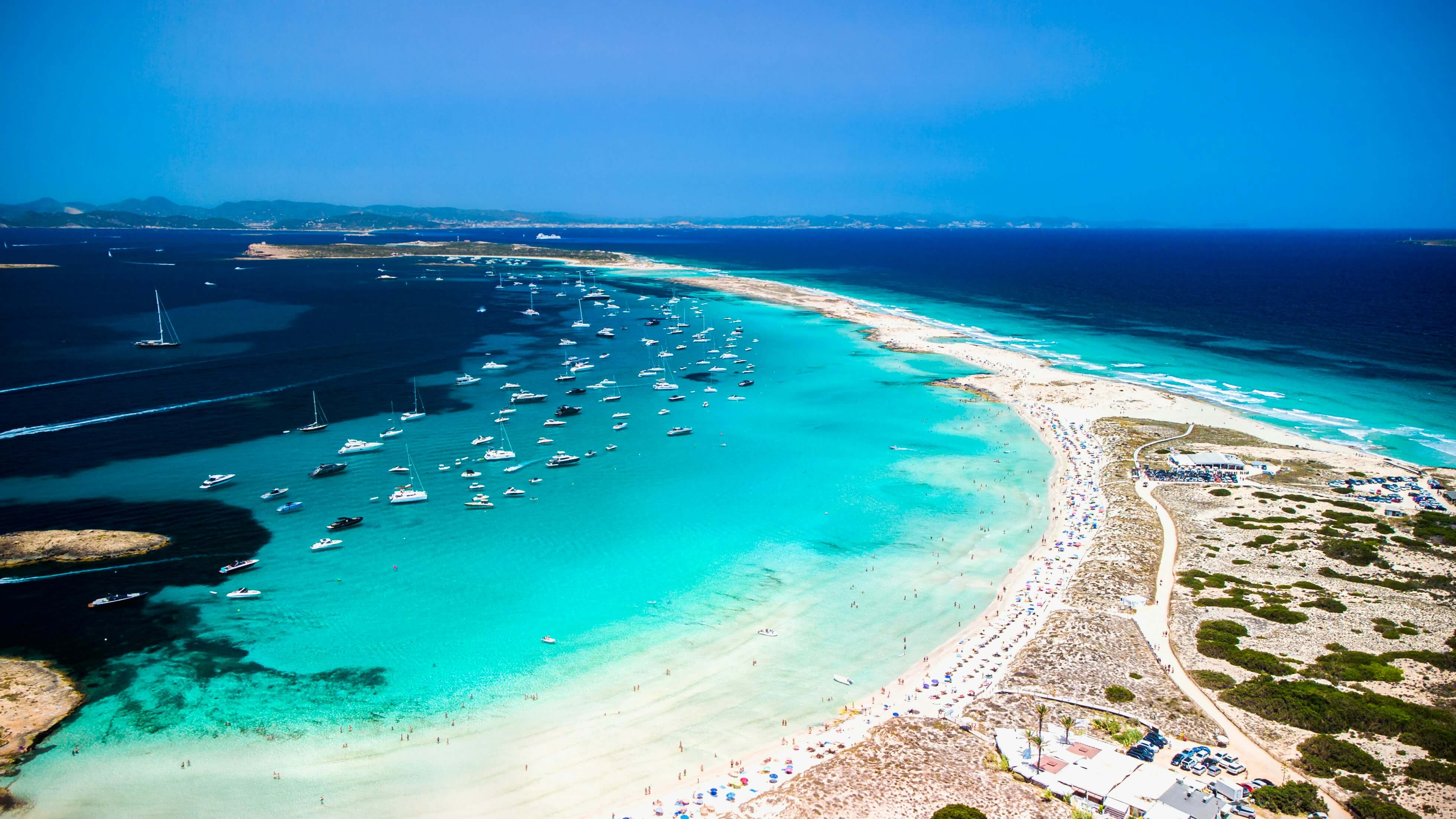 Aerial view of Ses Illetes beach in Formentera, Spain, with a long stretch of white sand surrounded by crystal-clear turquoise water, numerous boats anchored offshore, and people relaxing along the shoreline.