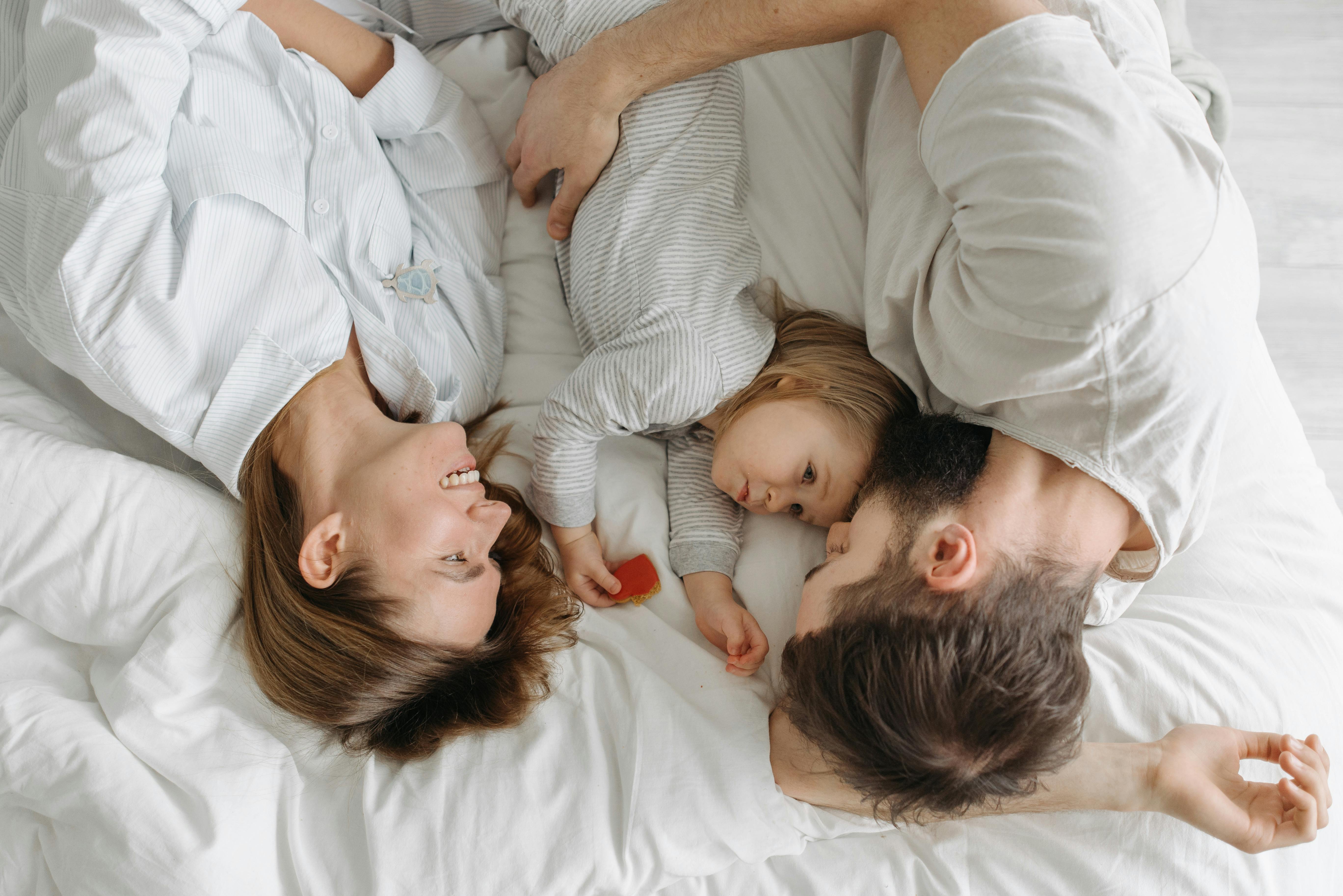 Mother, father, and young child lying together on a white bed, smiling and cuddling closely in a cosy family moment.