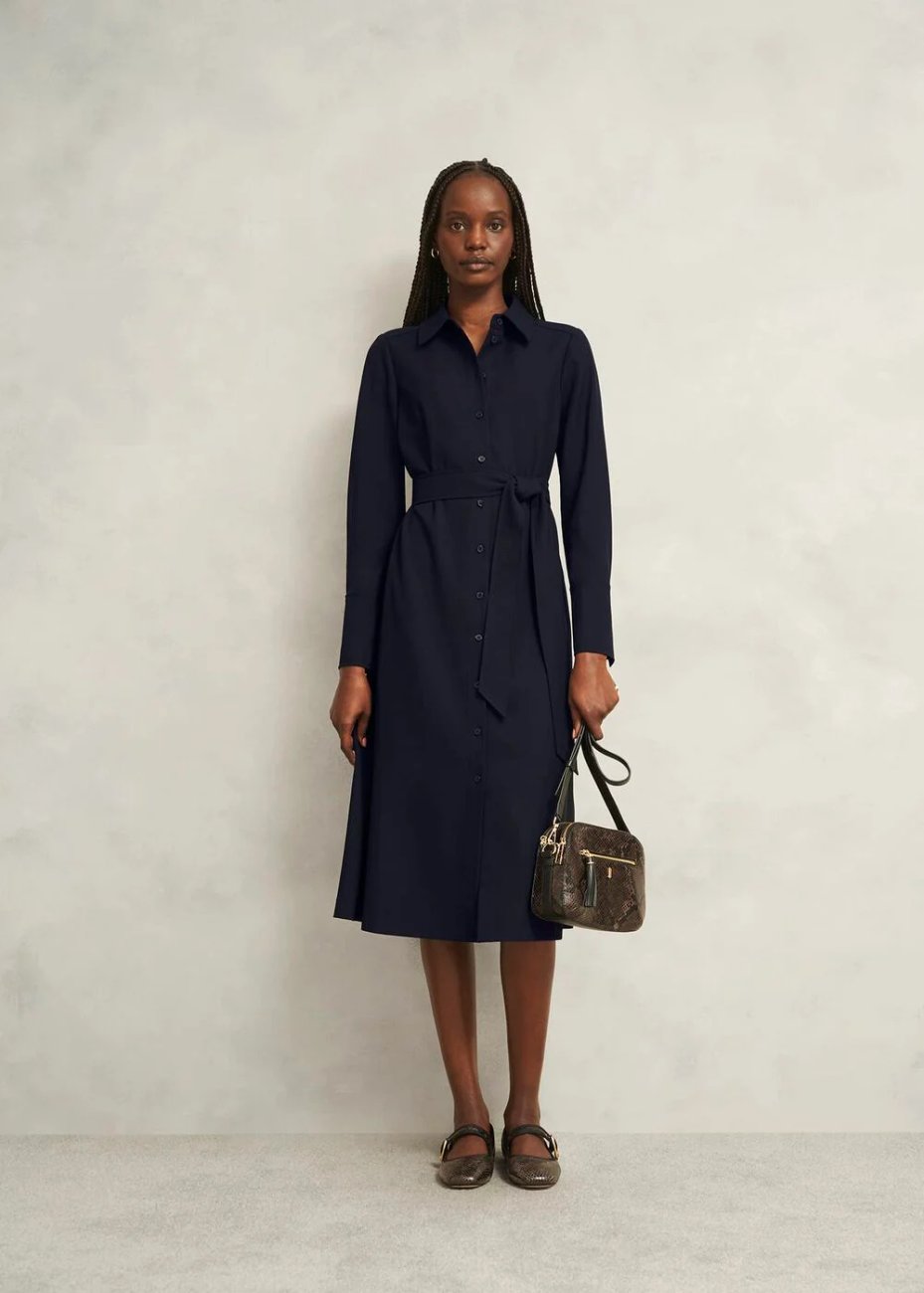 Model wearing Hobbs Lyla Dress in navy, a long-sleeve, tailored dress with a belt paired with a small handbag