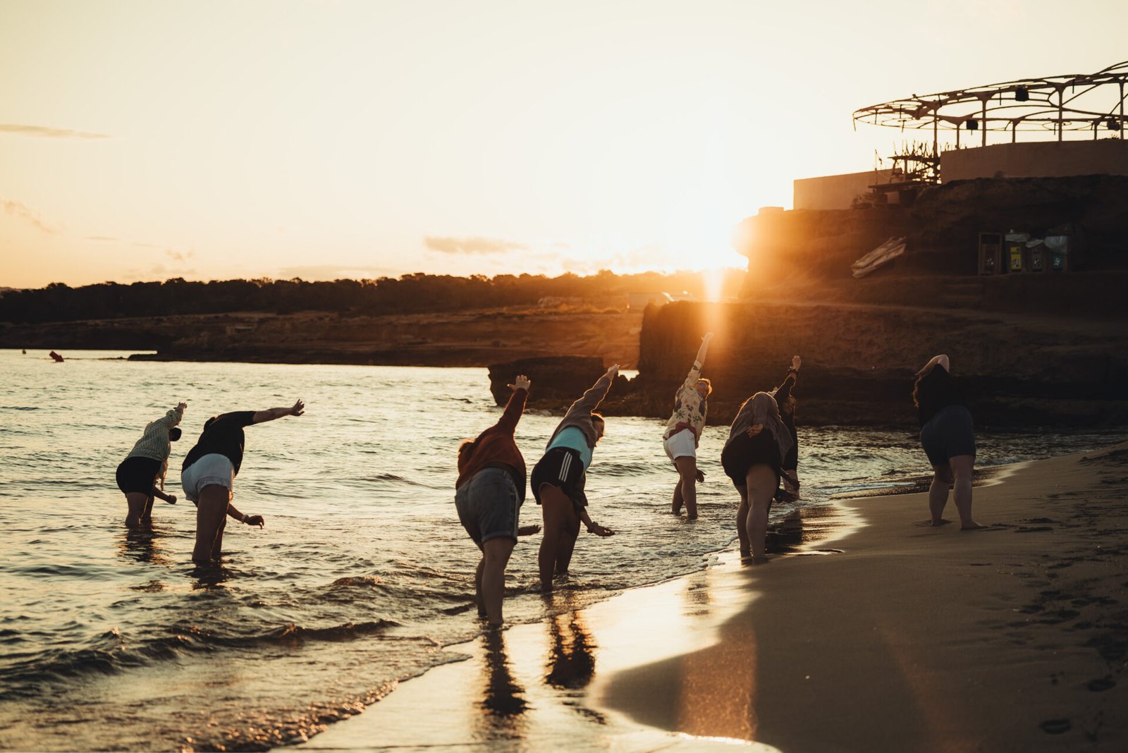At an Ibiza retreat a group of people standing in the shallow water at a beach during sunset, performing a synchronised side-stretching exercise with arms raised high