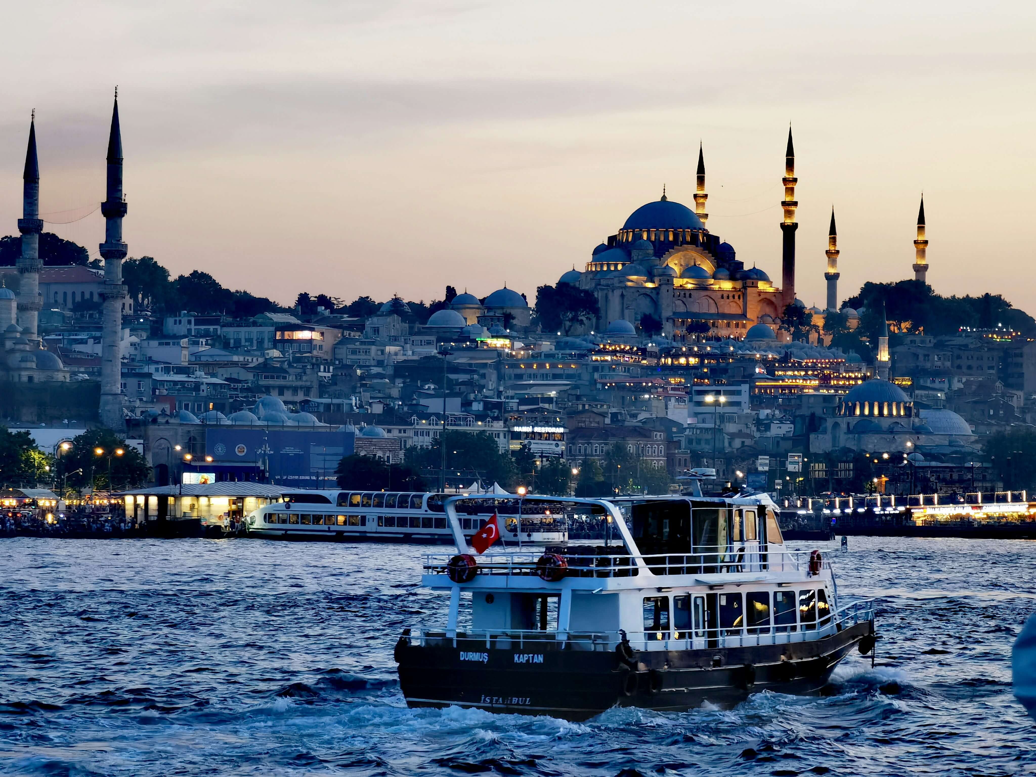 Passenger ferry sailing on the Bosphorus in Istanbul at sunset with the illuminated Hagia Sophia and mosque minarets rising above the city skyline in the background.