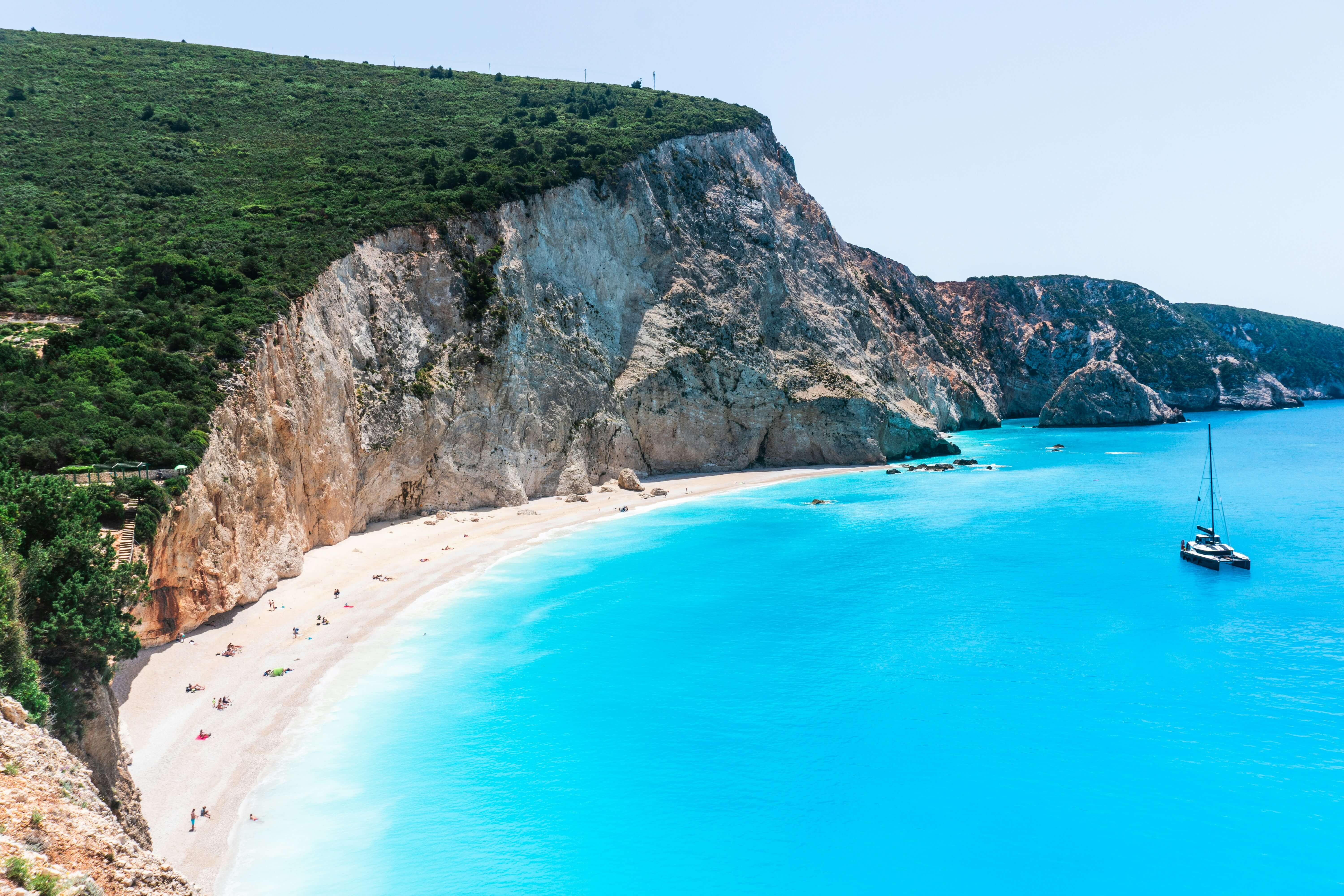 Secluded sandy beach beneath steep rocky cliffs in Lefkada Greece with vivid turquoise water and a lone sailboat anchored offshore