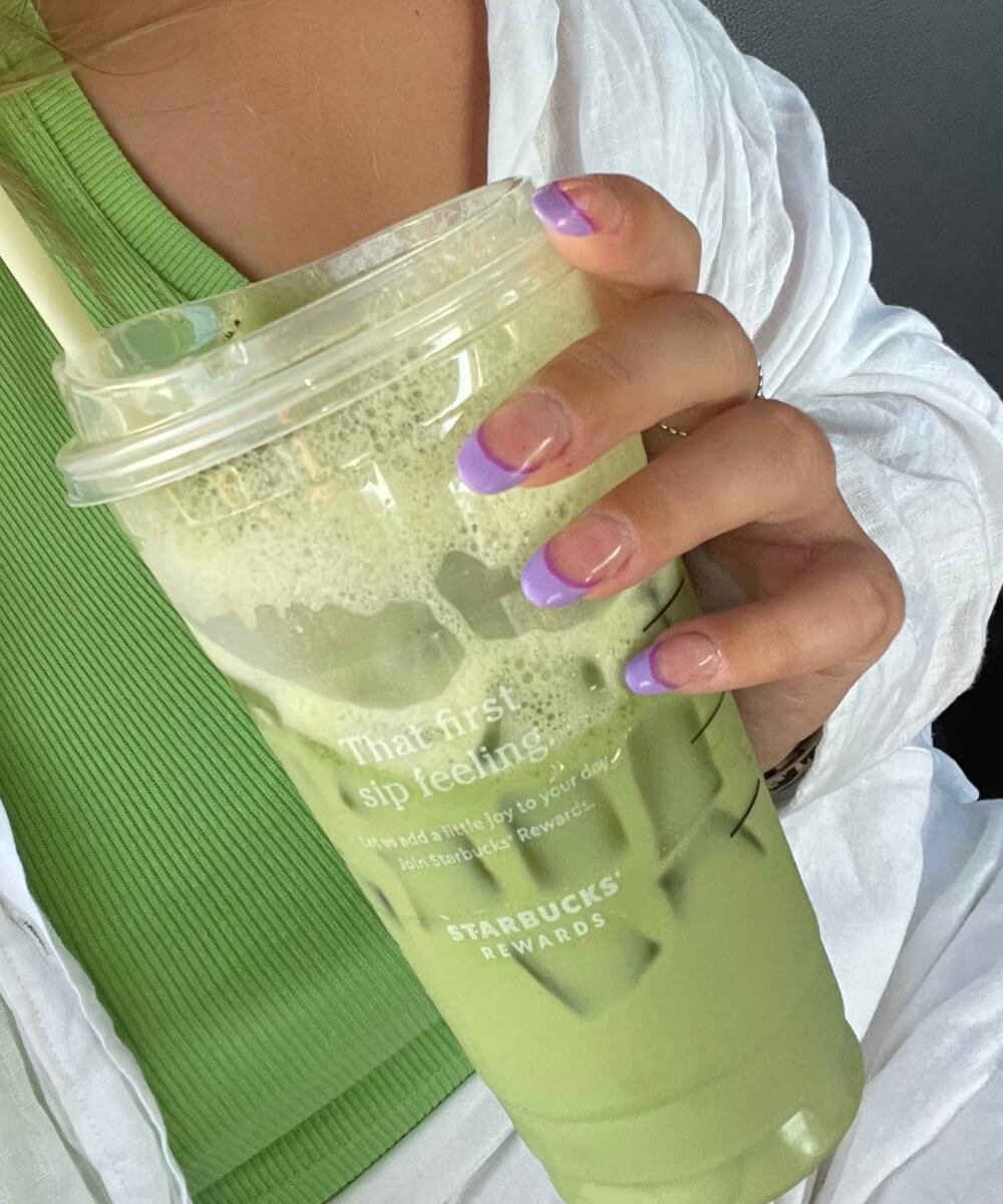 Close-up of almond-shaped nails with soft lilac French tips on a glossy nude base, holding an iced matcha latte, creating a clean and modern spring nail look