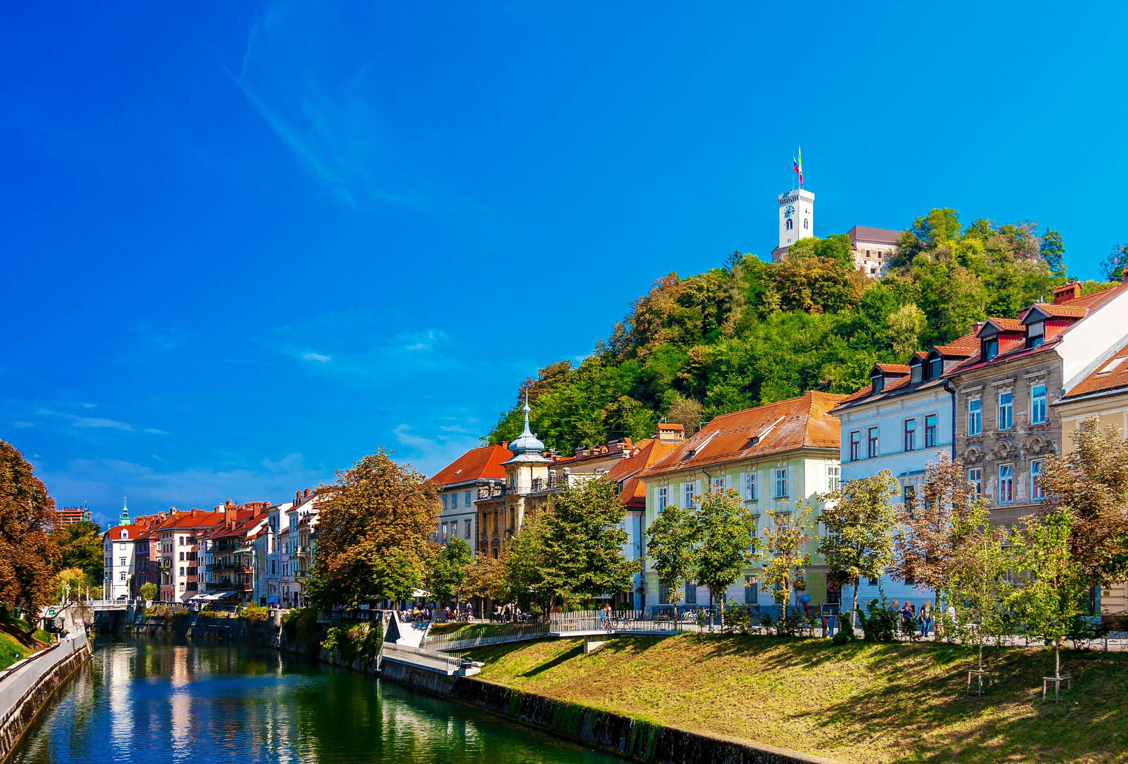 Colourful historic buildings line the Ljubljanica River in Ljubljana, Slovenia, with trees along the waterfront and Ljubljana Castle visible on a green hill under a clear blue sky.