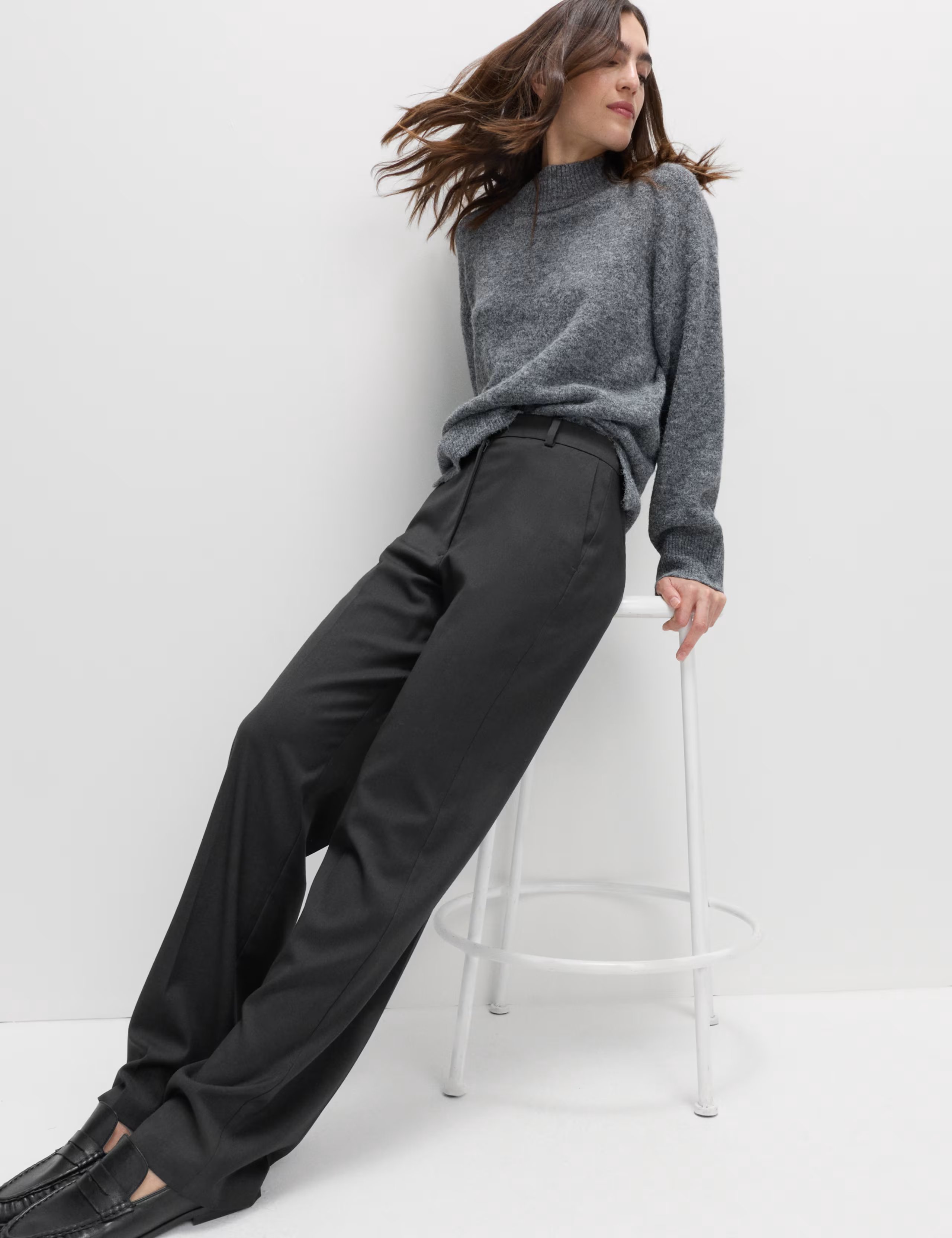 Model wearing M&S Straight Leg Trousers with Stretch in charcoal colour, leaning against a white high stool