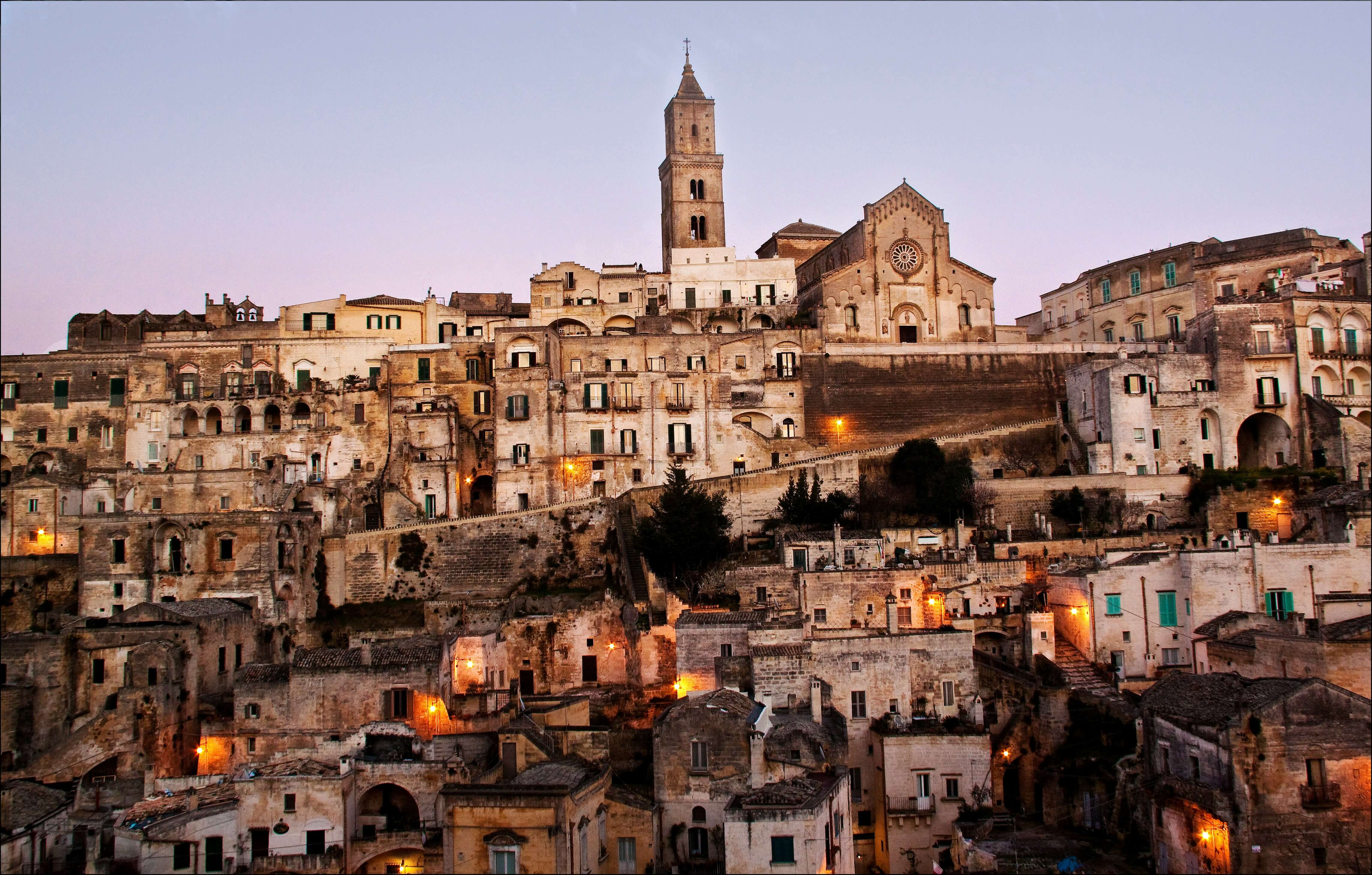 Historic stone buildings of the Sassi di Matera in Italy stacked along a hillside at dusk, with warm lights glowing from windows and a large cathedral and bell tower rising above the ancient city.