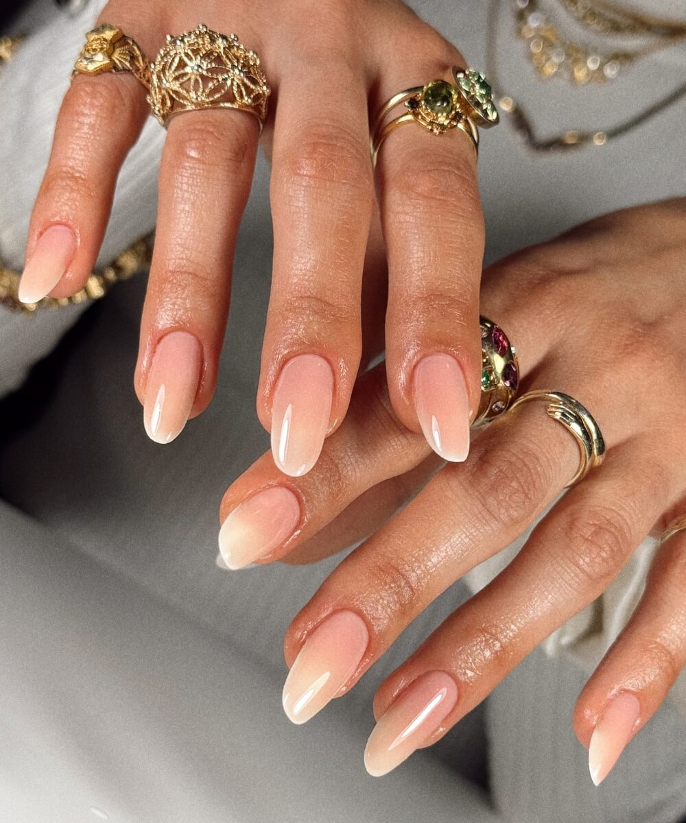 Close-up of almond-shaped nails with a soft milky pink to white ombré design and glossy finish, styled with gold jewellery for a clean, elegant spring nail look