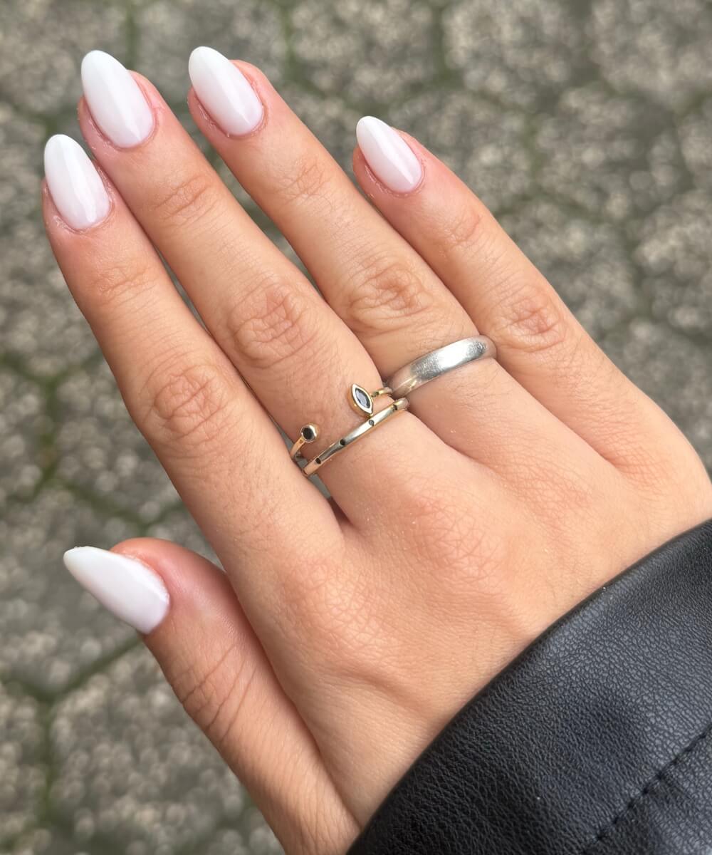Close-up of almond-shaped nails with a glossy milky white finish on a sheer base, styled with delicate gold and silver rings for a minimal spring nail look
