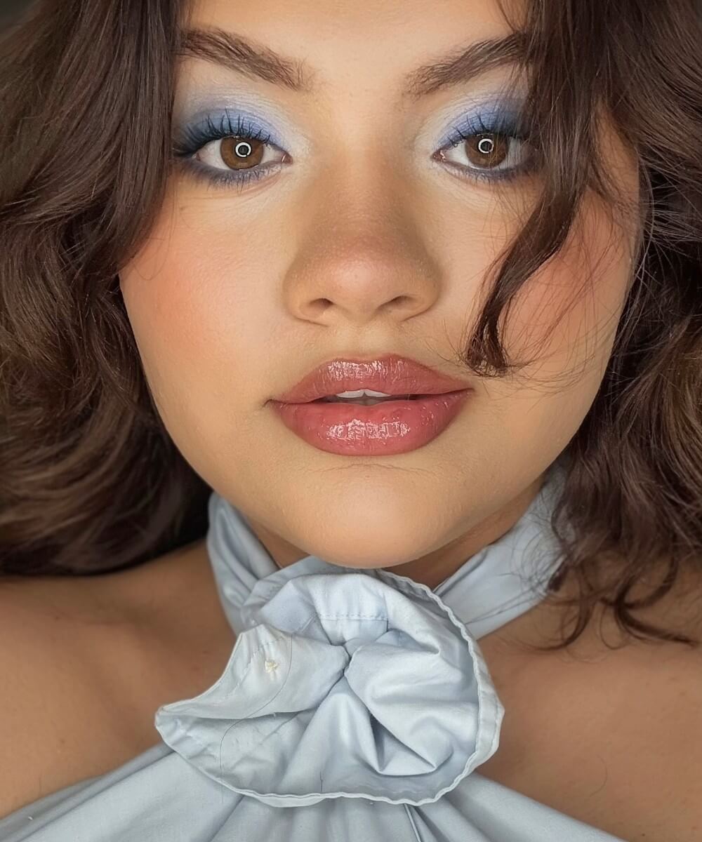 Close-up of a person with blue eyeshadow and glossy lips, wearing a light blue top with a bow