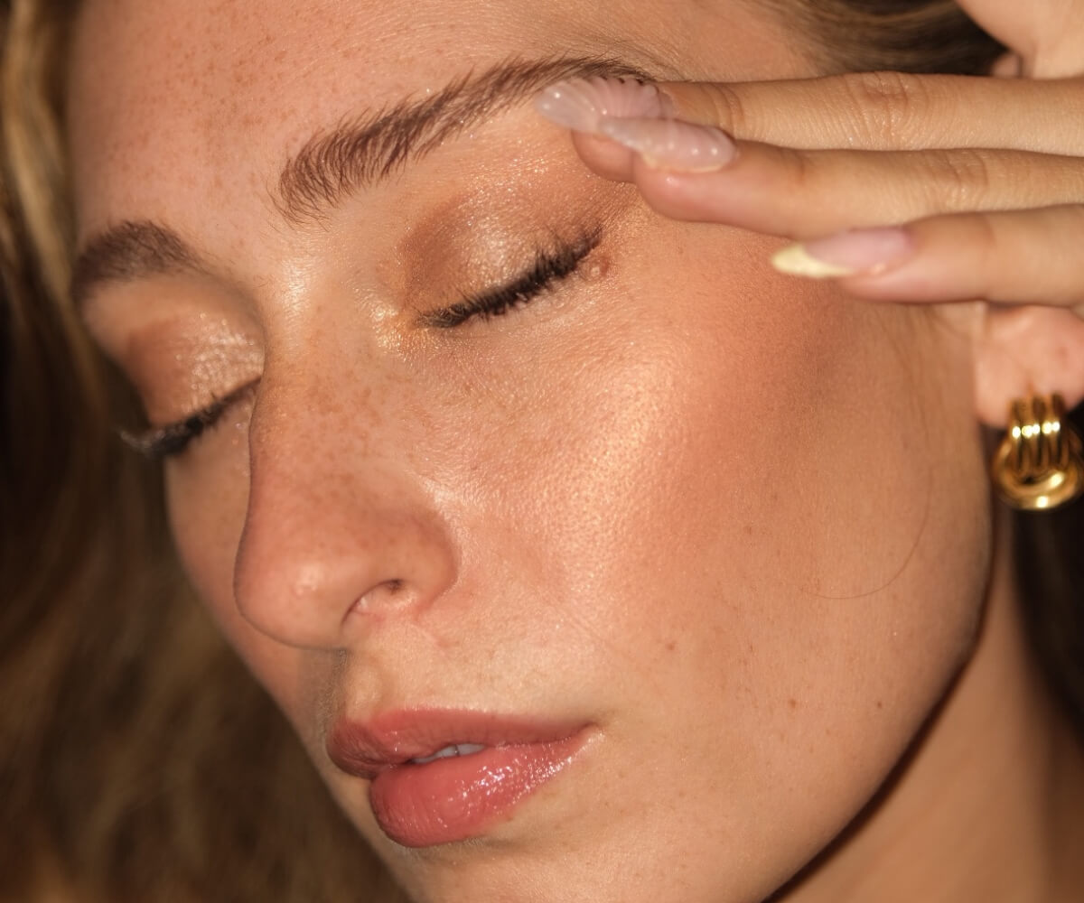 Close-up of a woman with natural glowing skin applying skincare near her eyebrow, showing softly defined, natural-looking semi-permanent eyebrows