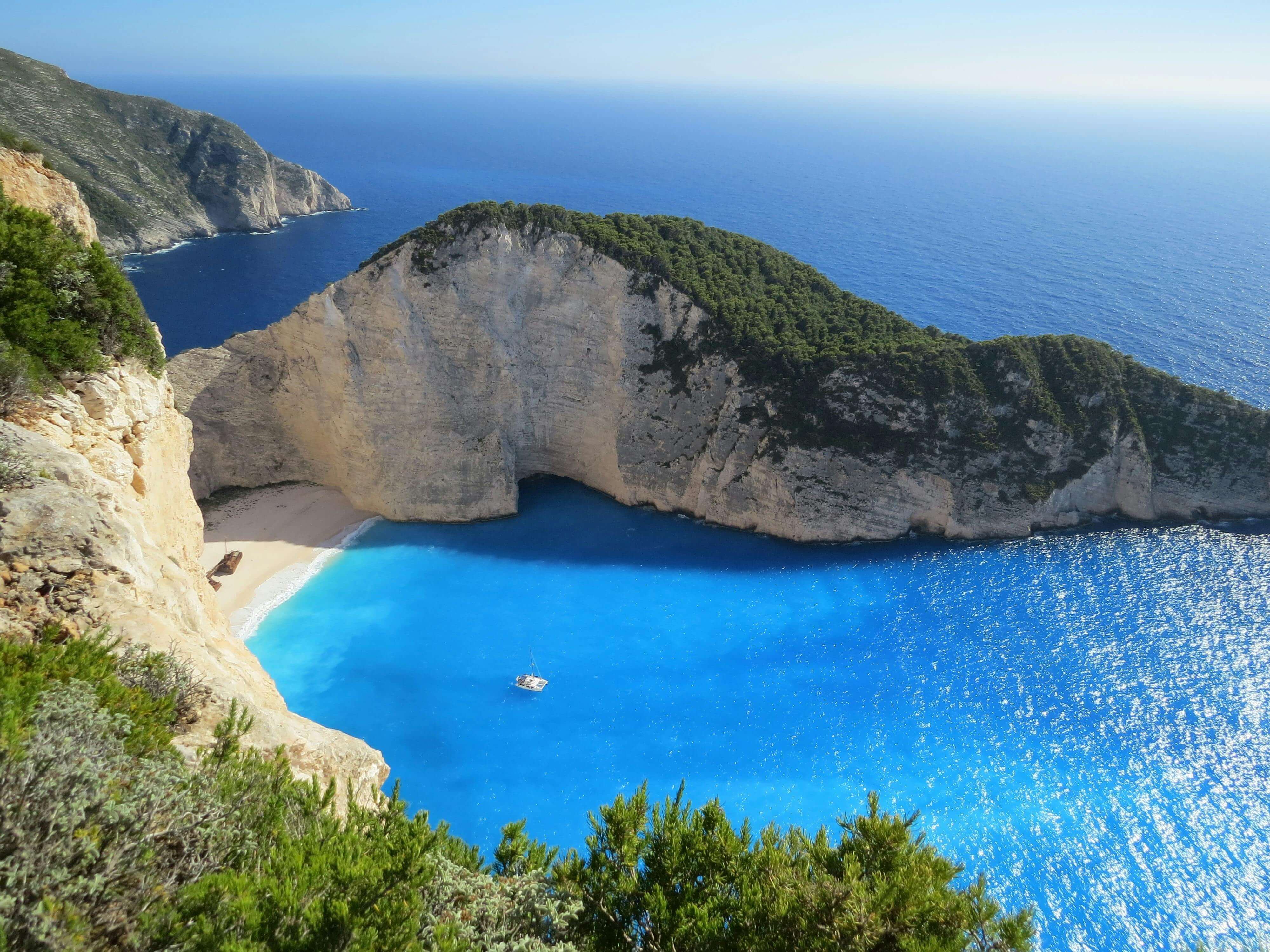 Aerial view of Navagio Beach in Zakynthos, Greece, featuring a secluded white-sand cove surrounded by towering limestone cliffs and vivid turquoise water, with a shipwreck resting on the beach and a small boat floating in the bay.