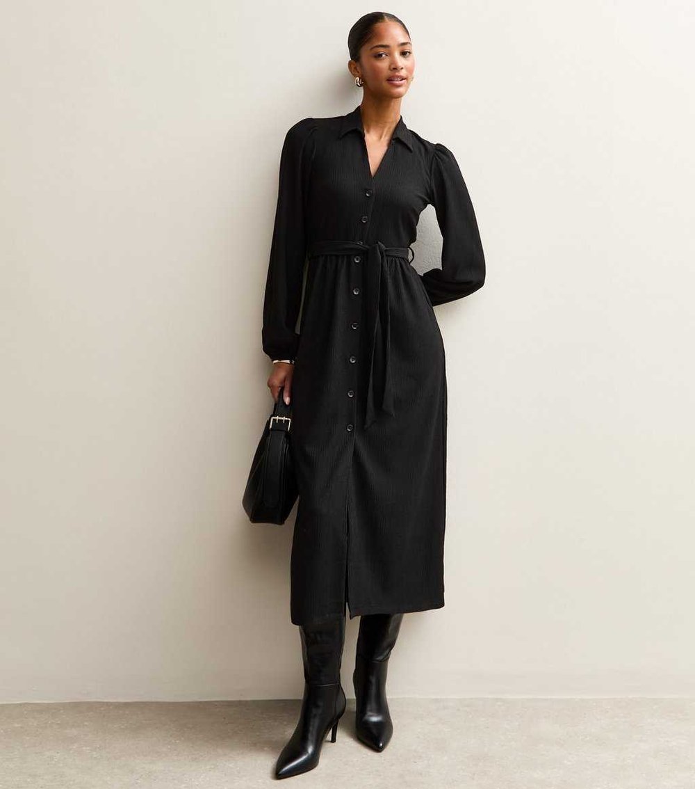 Model wearing New Look black crinkled shirt dress with long sleeves, standing against a white wall, holding a black handbag and wearing black high heels