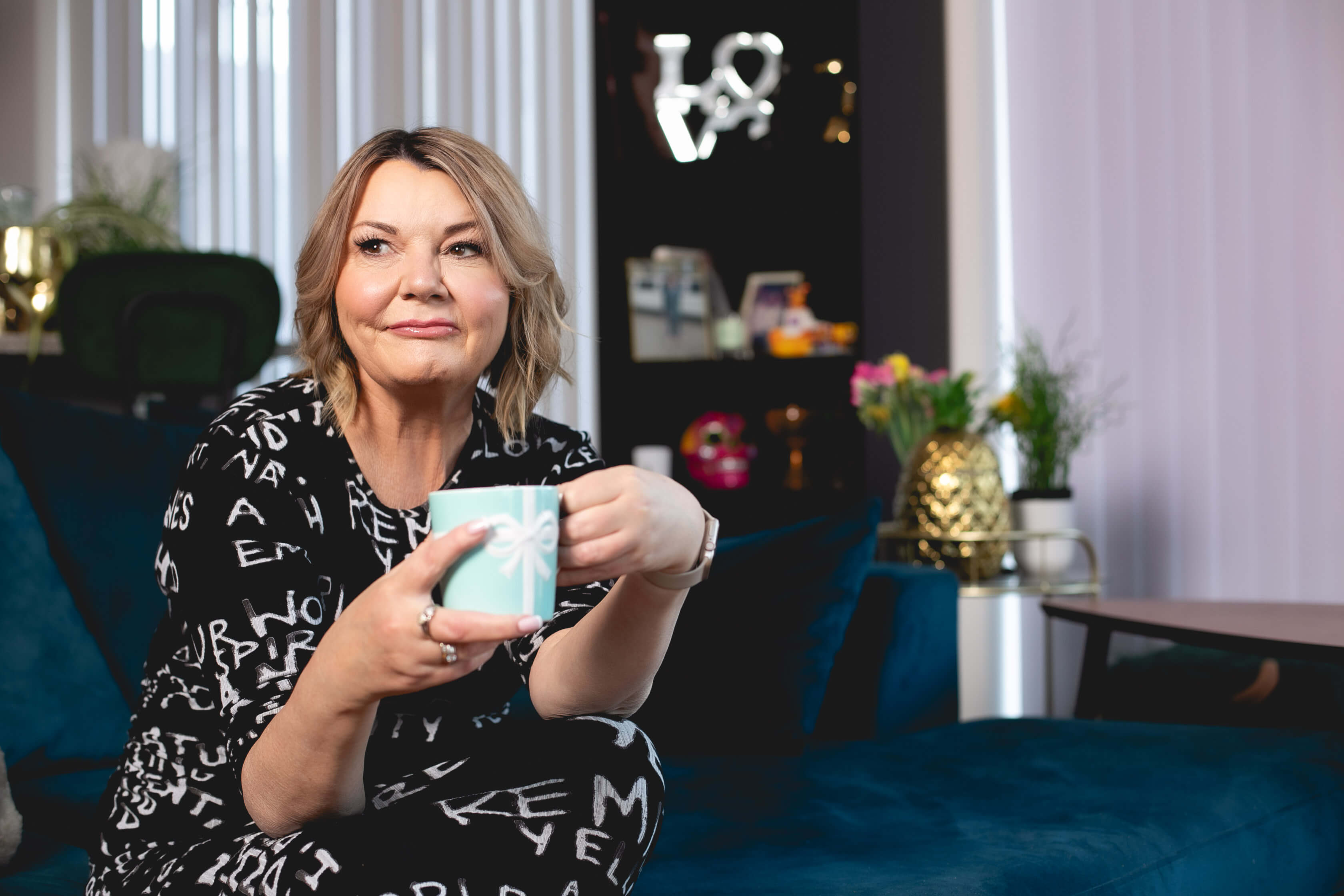 Nicky Wake wearing a black patterned dress sitting on a sofa holding a mug after quitting alcohol and embracing sober life - midlife sobriety