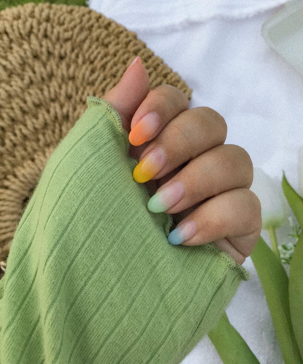 Close-up of almond-shaped nails with soft pastel ombré coloured french tips in rainbow shades including orange, yellow, green and blue, creating a fresh and colourful spring nail look
