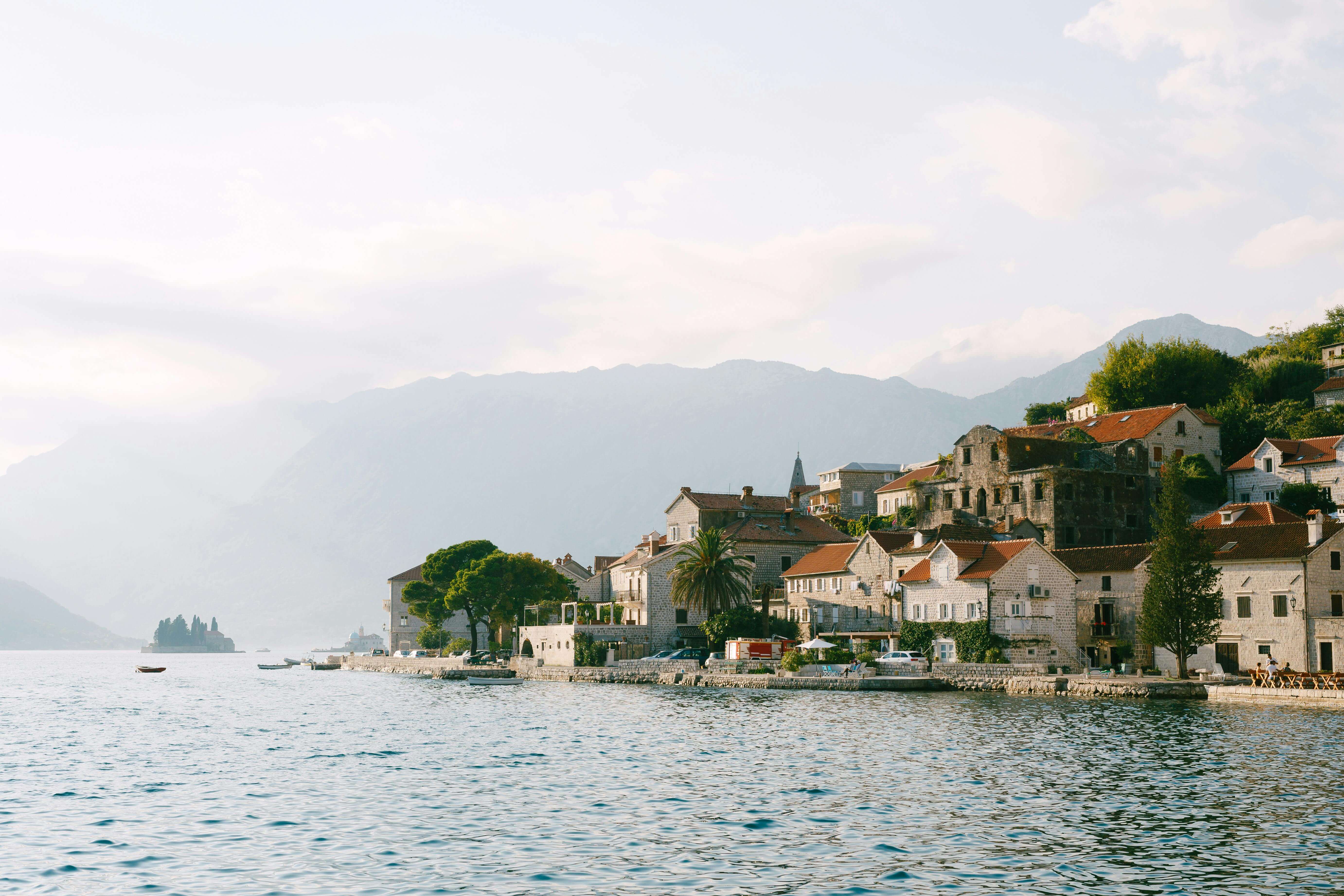 Scenic view of Perast, Montenegro, with historic stone houses and red-tiled roofs along the waterfront of the Bay of Kotor, calm water in the foreground, and misty mountains rising in the background.
