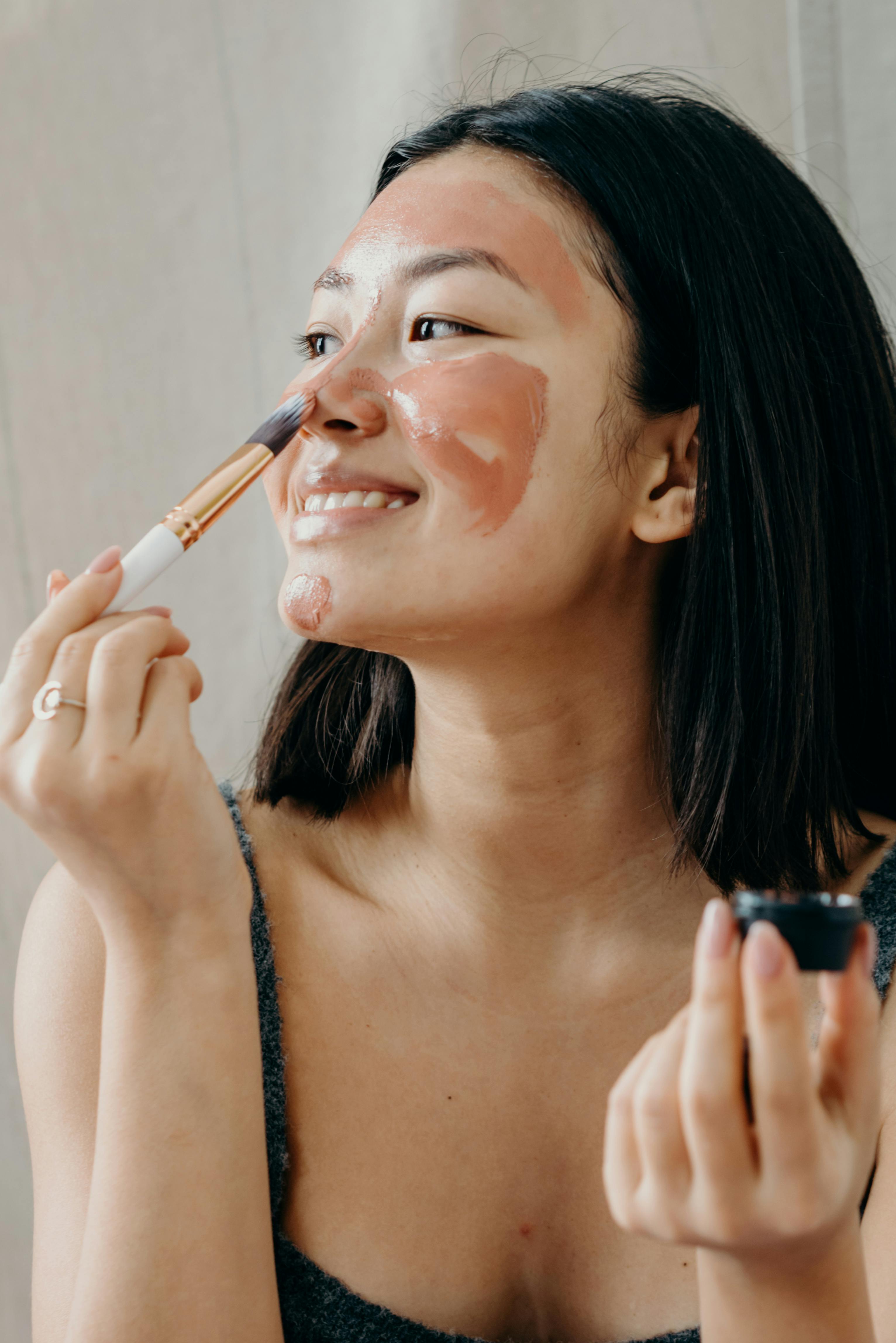A smiling Korean woman applies a face mask using a make-up brush.