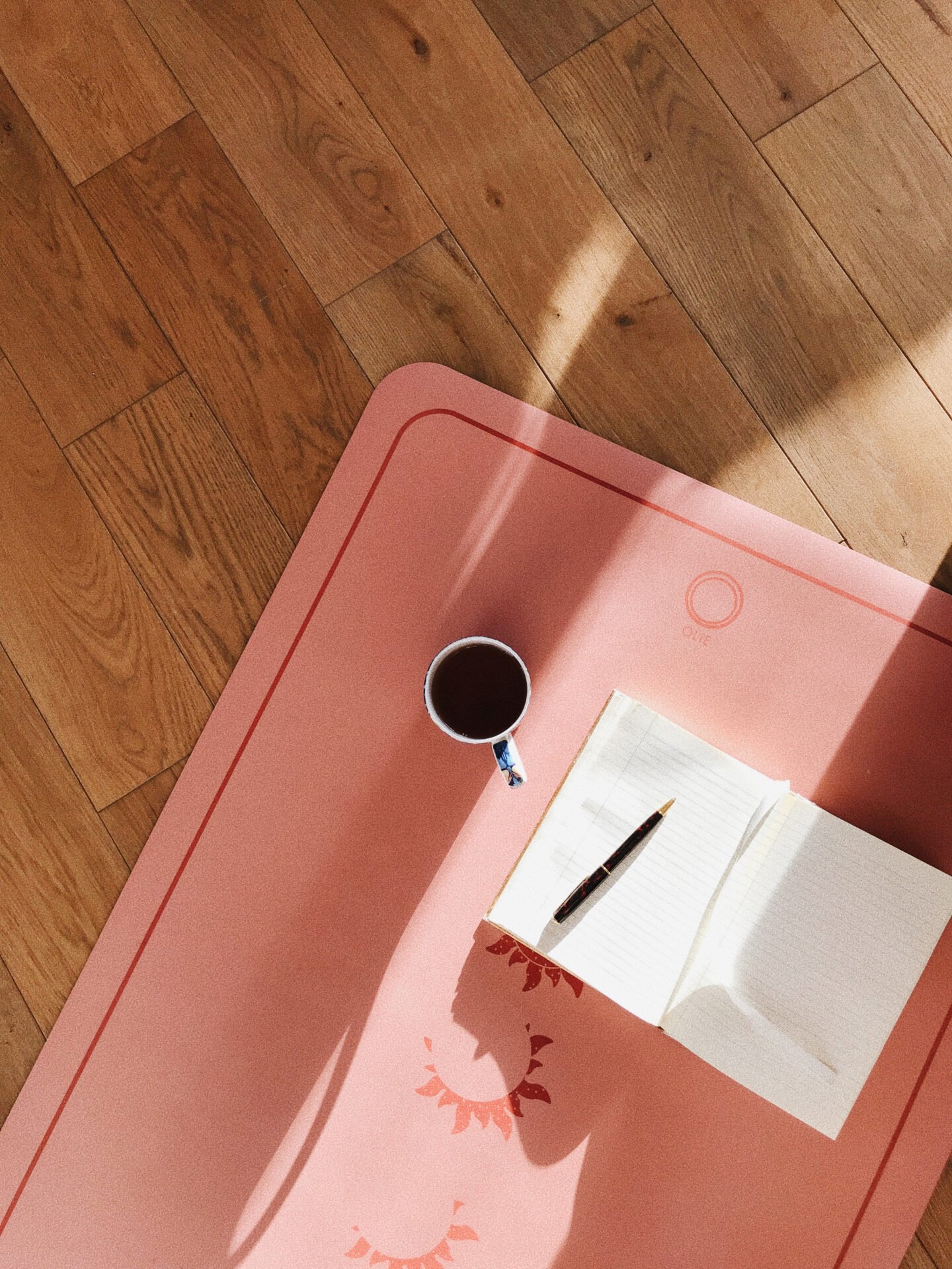 A pink yoga mat with sun motifs on a wooden floor, featuring an open wellness journal, a pen, and a cup of coffee under natural sunlight from a window representing the idea of nervous system regulation techniques