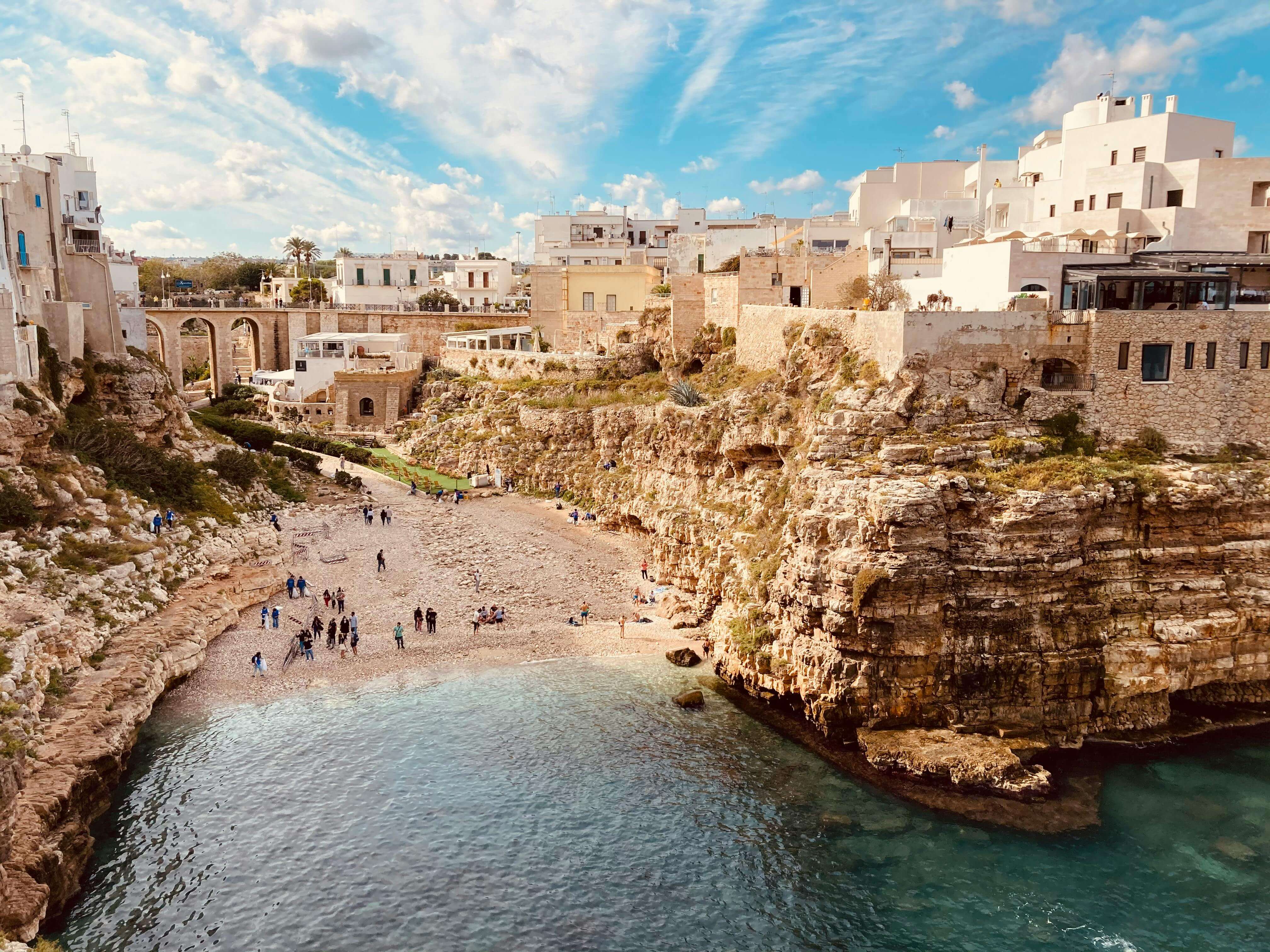 View of a rocky cove with turquoise water and a small pebble beach surrounded by dramatic limestone cliffs, with people walking along the shore and white buildings and a stone bridge overlooking the seaside town above.