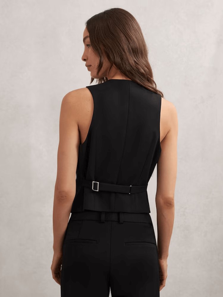 A back view of Gabi Single-Breasted Twill Waistcoat in Black, a fitted black vest and pants. The vest has a belt detail