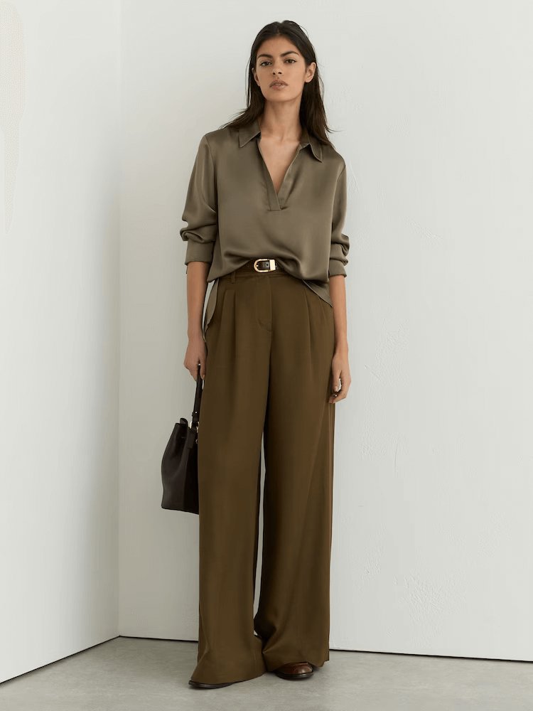 Model wearing Reiss Marina Silk Open Collar blouse in khaki green with a classic collar, long sleeves, and a V-neck, styled with brown trousers and a small, black bag