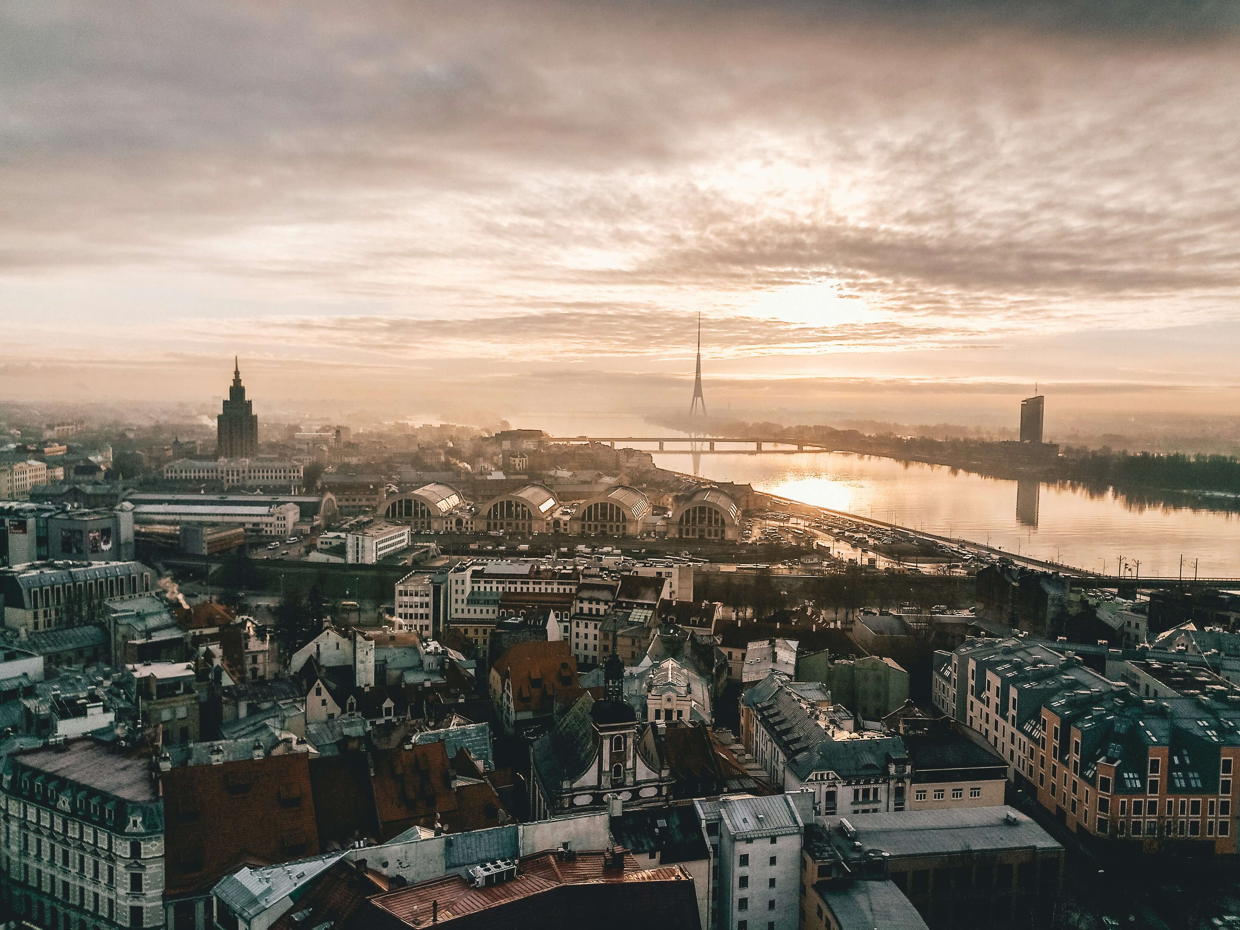 Aerial view of Riga, Latvia at sunrise with the Daugava River reflecting warm light, the Riga TV Tower in the distance, bridges crossing the river, and historic and modern buildings spread across the city.