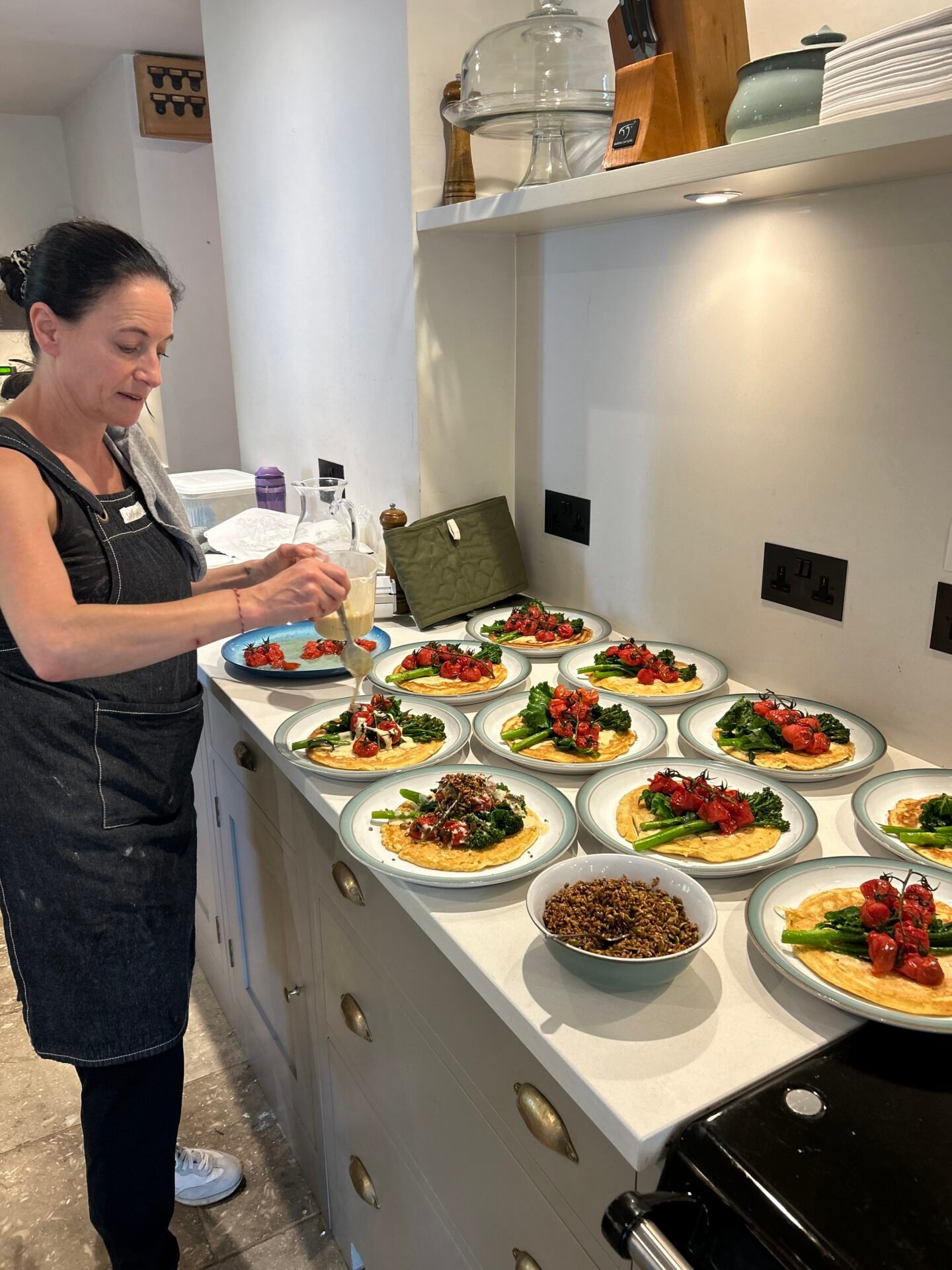 At Rise & Revive women-only retreat, a woman in a dark apron drizzles sauce onto plates of crepes topped with roasted tomatoes and greens arranged on a kitchen counter for service