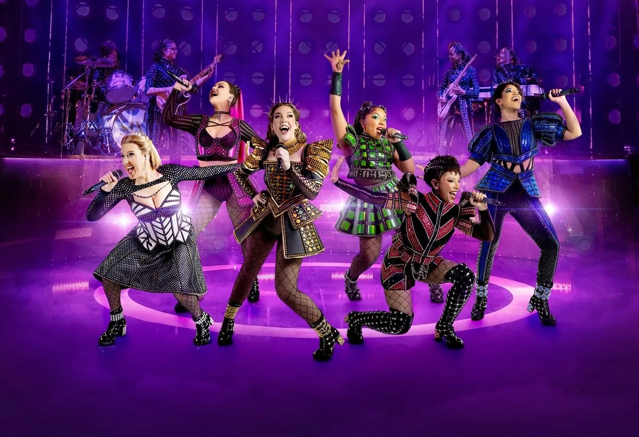 six women with microphones in hands singing on a purple stage in six the musical show