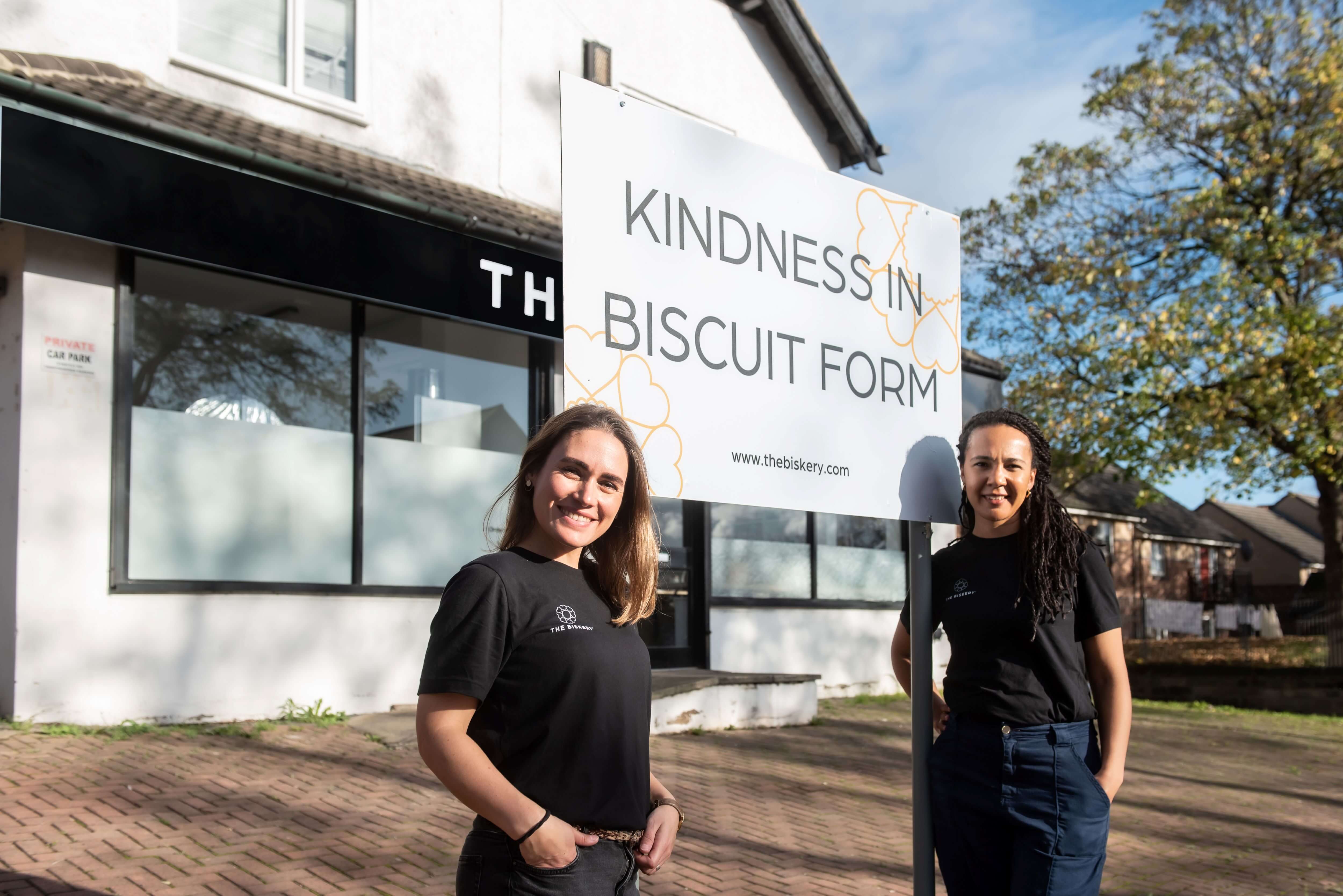 The Biskery co-founders Lisa de Boer and Saskia Roskam standing outside The Biskery headquarters in Leeds beside a 'Kindness in Biscuit Form' sign