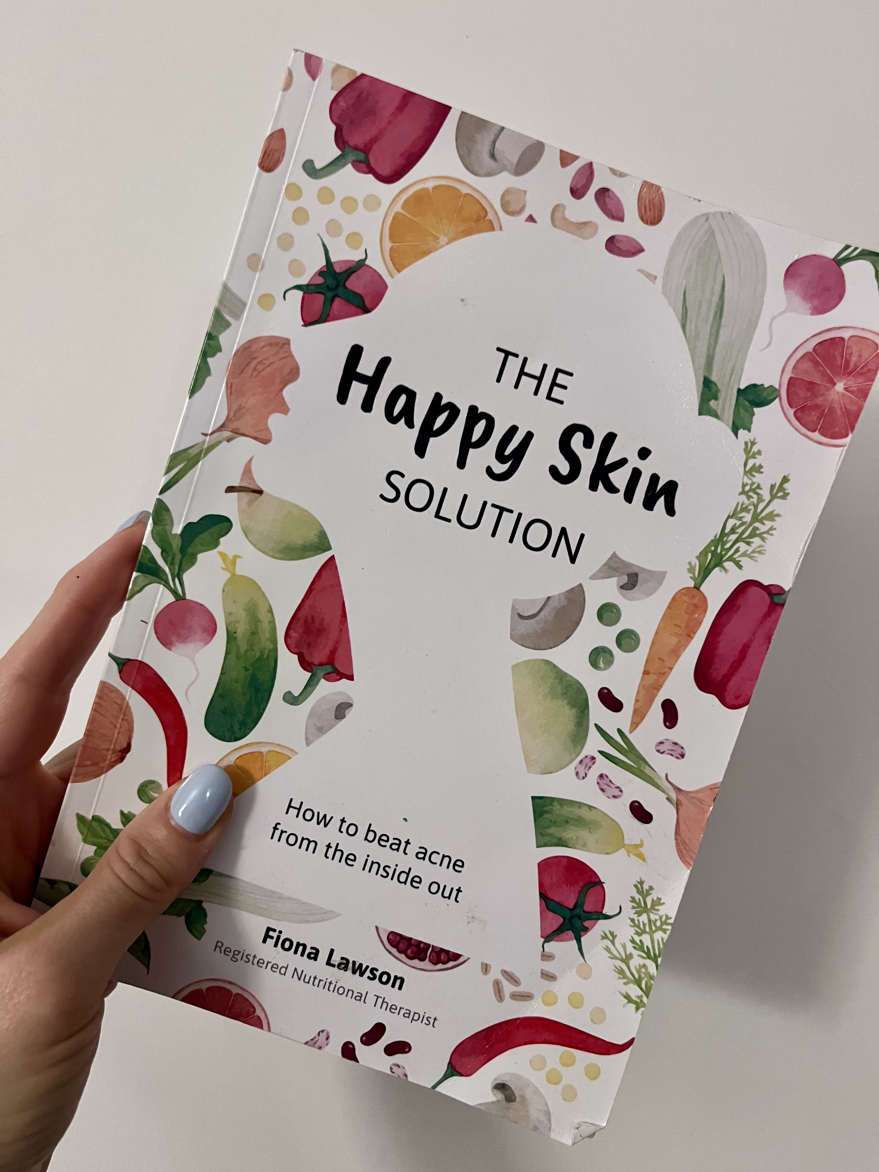 A woman's hand holding up a book called The Happy Skin Solution - How to beat acne from the inside out by Fiona Lawson