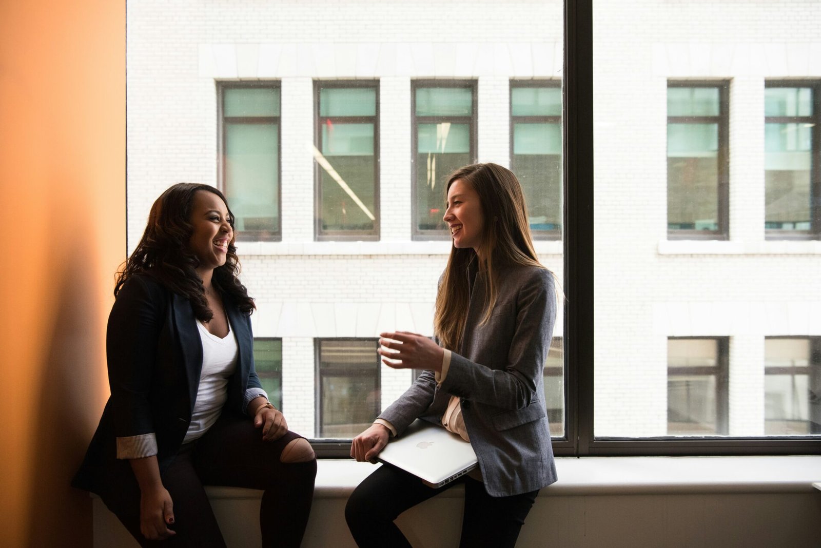Two businesswomen engaged in conversation, standing in front of a large window with natural light streaming in - best investors for women businesses