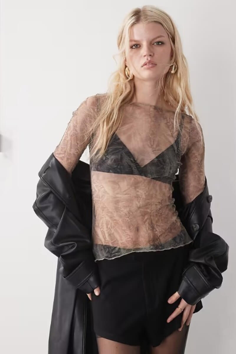 A model wearing Weekday sheer mesh long sleeve top over a black bra, paired with a black leather jacket and shorts
