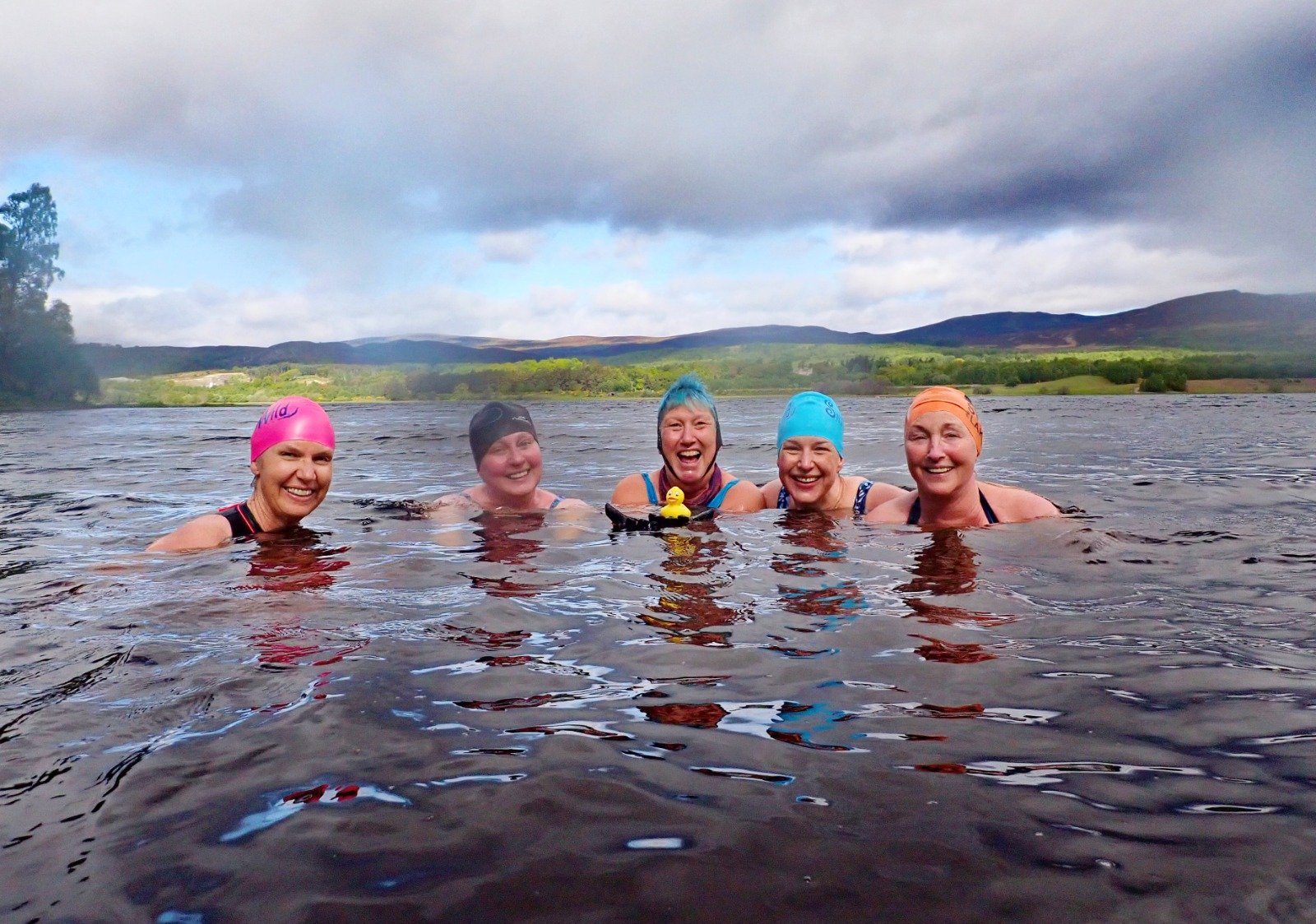 Five women wearing colourful silicone swim caps swimming together in a dark lake with mountains and a cloudy sky in the background on a bright day at The Wilderness Retreat - best women-only retreats